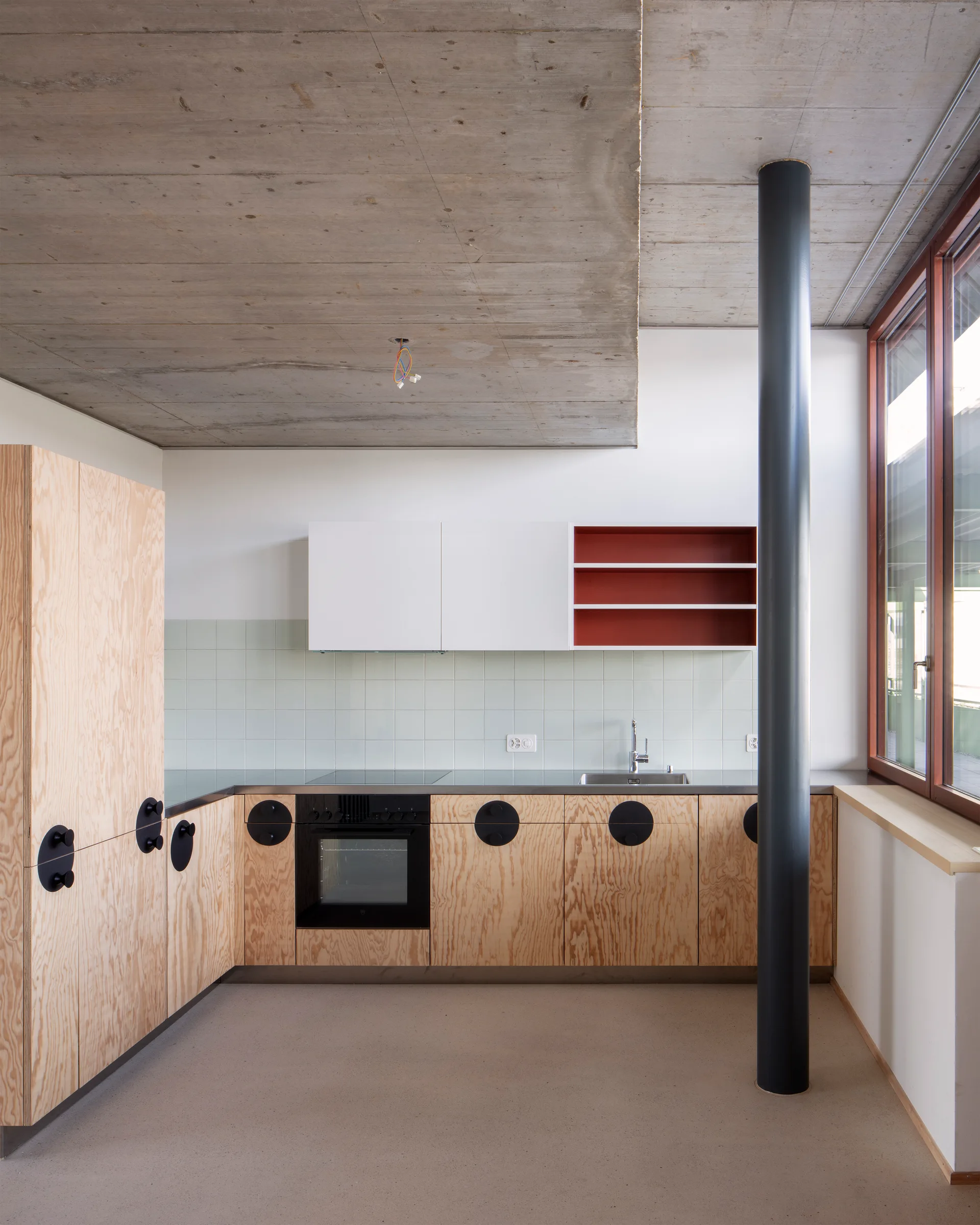 A minimalist kitchen featuring plywood base cabinets with large round black handles, a light green tile backsplash, and an exposed concrete ceiling.
