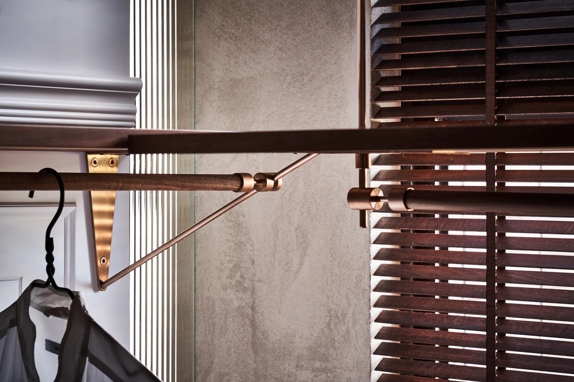 A close-up of a custom closet system featuring a dark wood clothing rod, brass brackets, a wood shelf, and slatted window blinds.