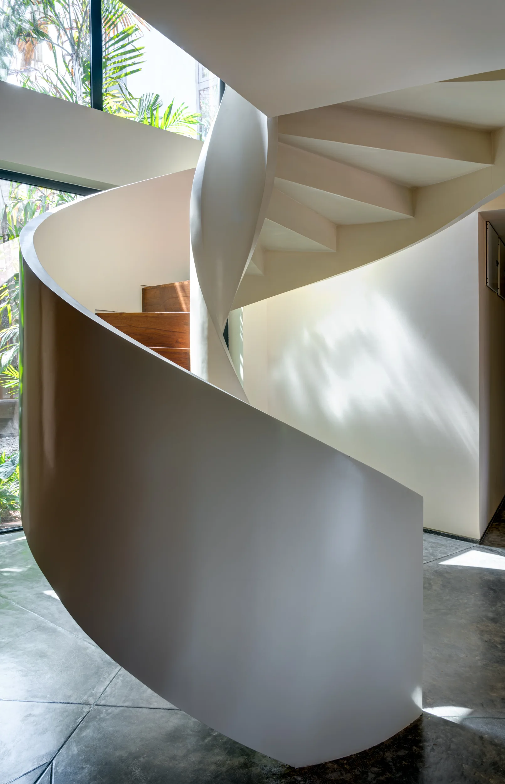 A sculptural white spiral staircase with dark wood treads sits on a polished concrete floor next to a window.