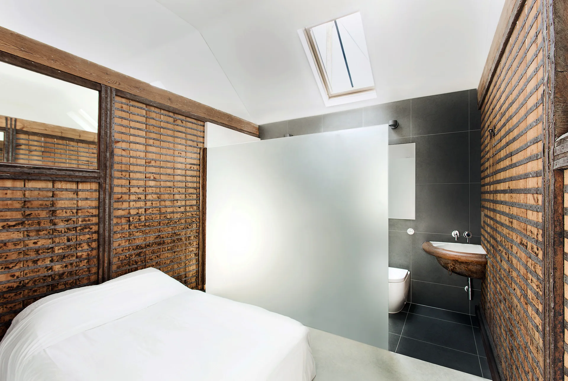A rustic loft bedroom with weathered wood walls features a bed separated from a grey tiled bathroom by a large frosted glass partition.