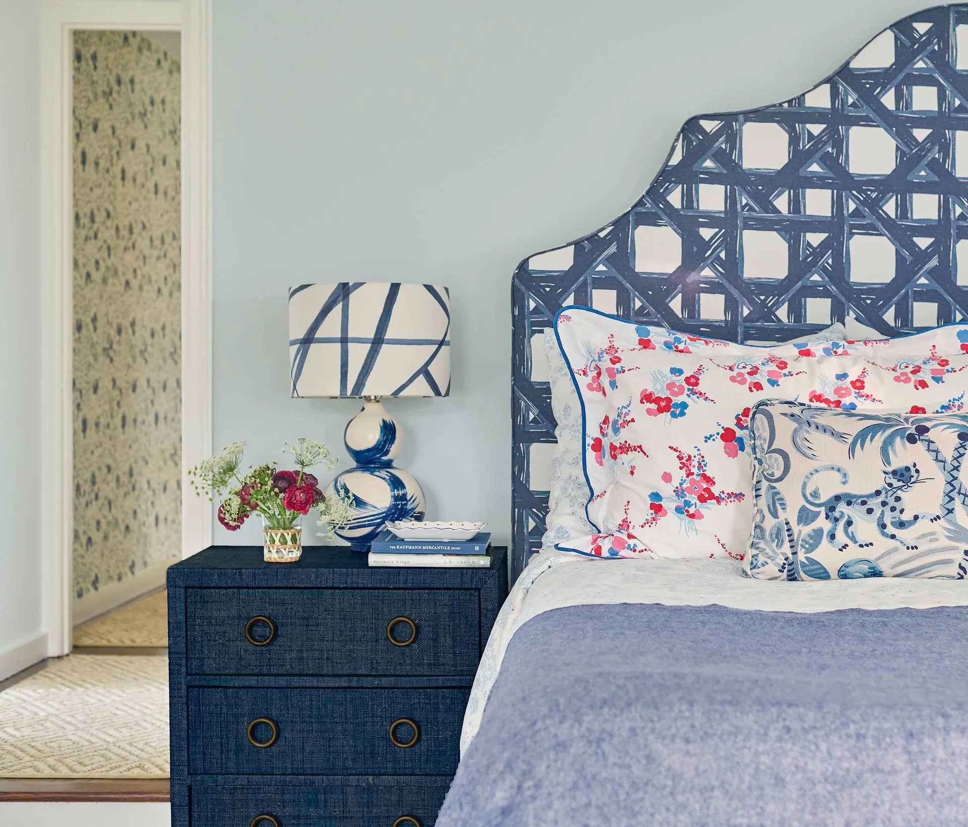 A bedroom scene features a curved blue patterned headboard next to a navy blue textured nightstand with a ceramic lamp and colorful pillows.