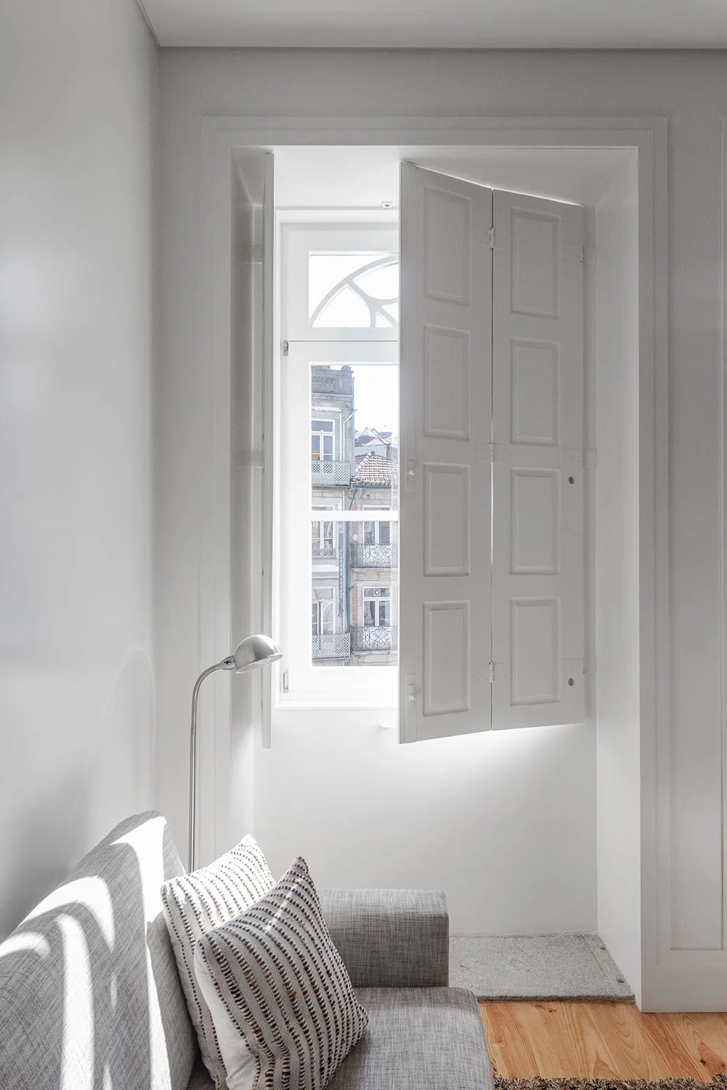 A gray fabric sofa with patterned pillows sits next to a tall window with white paneled interior folding shutters.