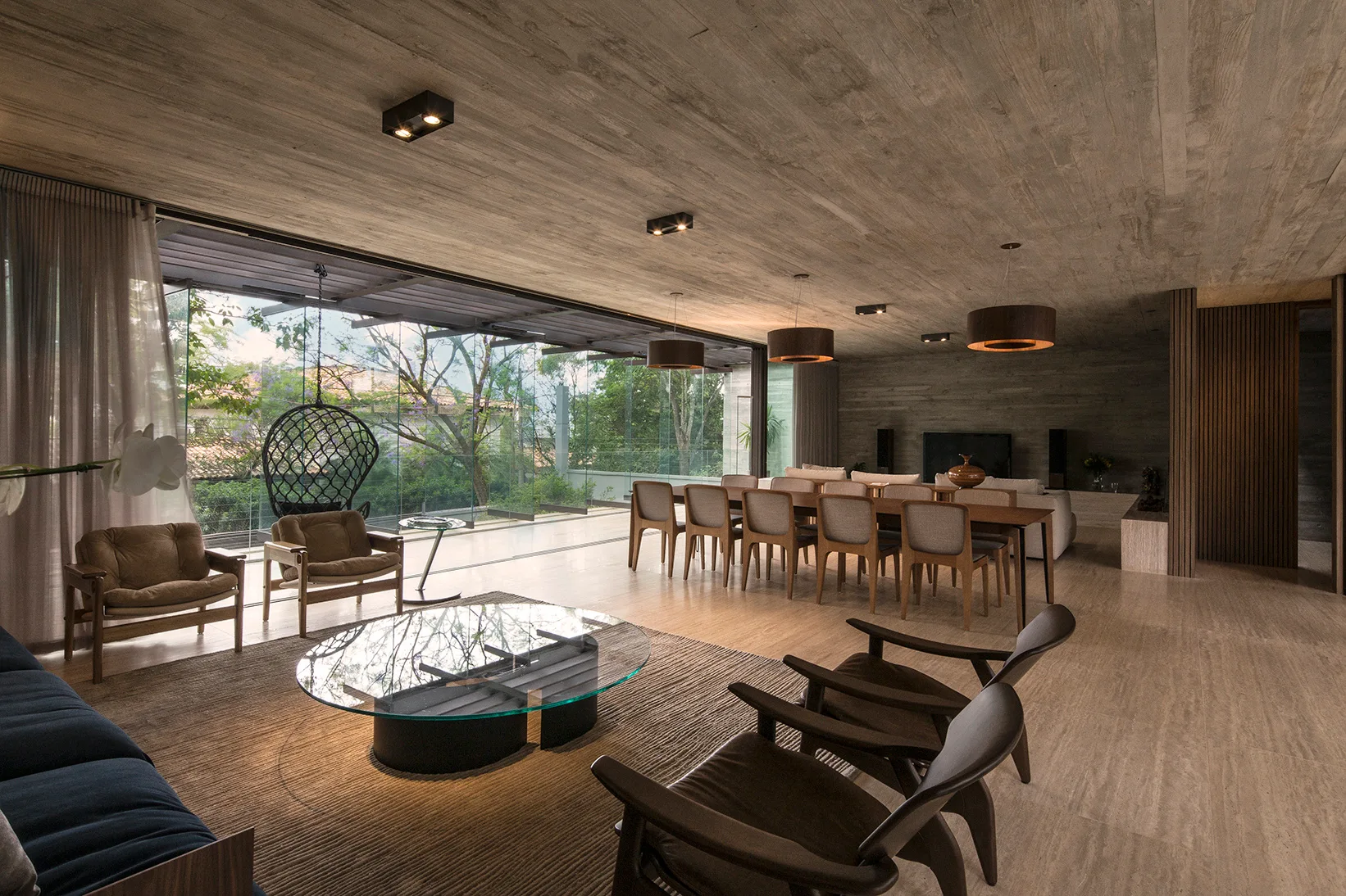 A spacious open-plan living room with a textured concrete ceiling, large glass walls, a wooden dining table, and a glass-topped coffee table.