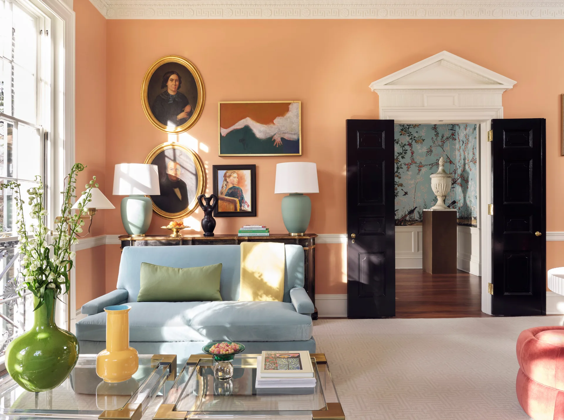 A pastel living room features peach walls, a light blue sofa, and high-gloss black double doors opening to an adjacent wallpapered space.