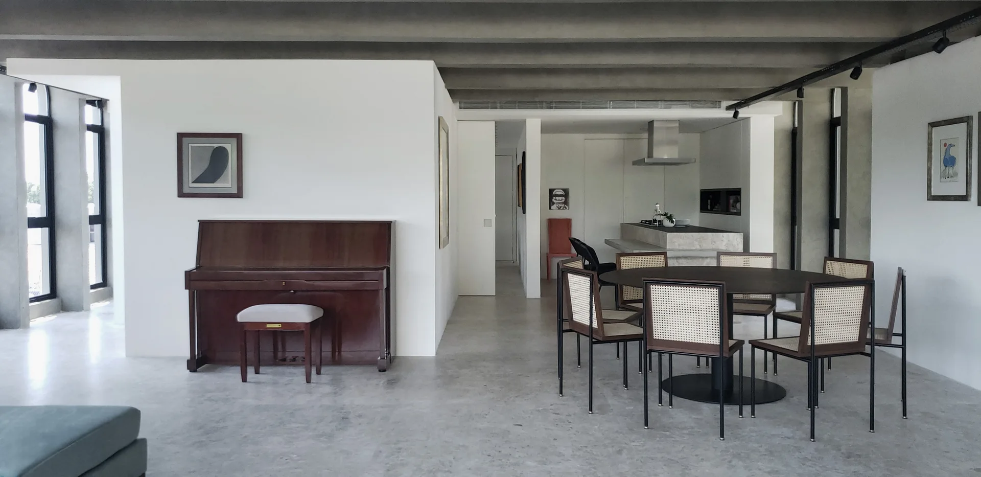 An open-plan room features a round black dining table with woven cane chairs, a wooden upright piano, concrete floors, and exposed ceiling beams.
