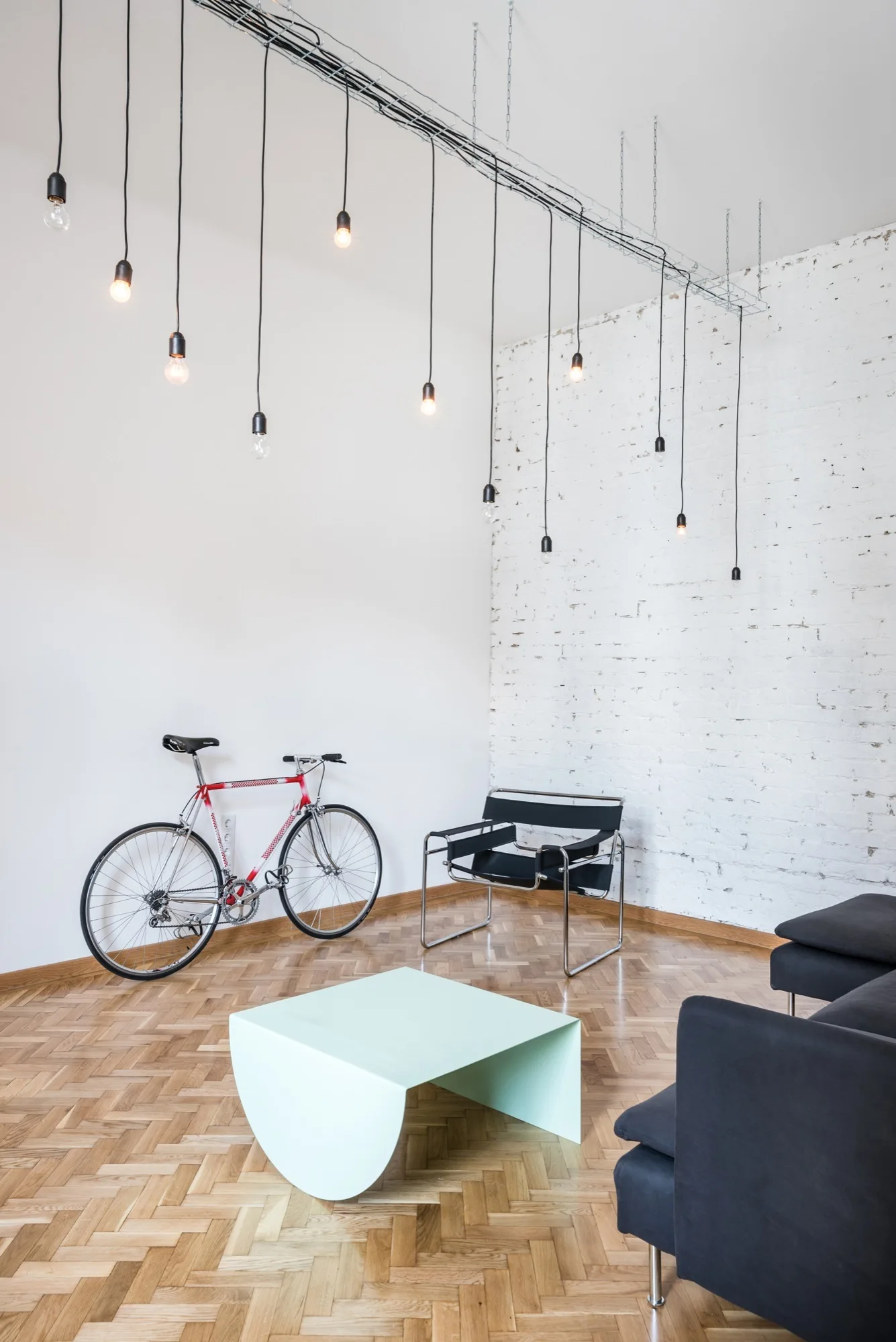 A minimalist living room featuring a herringbone wood floor, a white painted brick wall, a black tubular armchair, and hanging pendant lights.