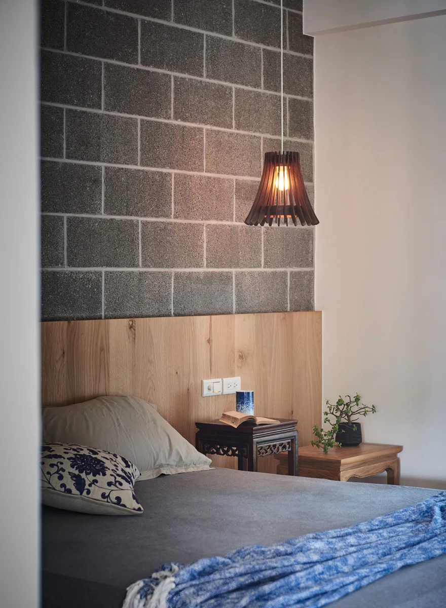 A bed with grey bedding and a blue throw blanket is positioned against a cinder block feature wall with a light wood headboard panel.