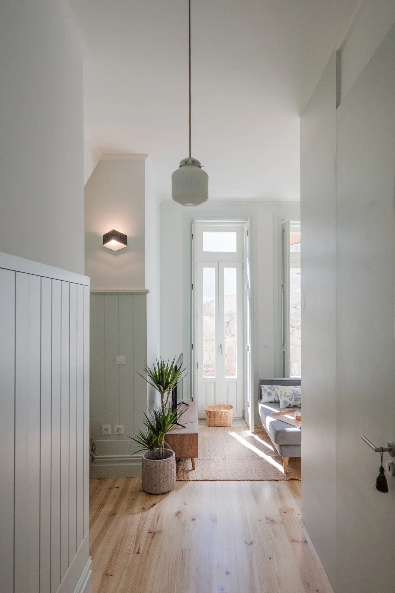 A bright hallway leading into a living room with pale green wainscoting, light wood floors, a grey sofa, and a tall French door.