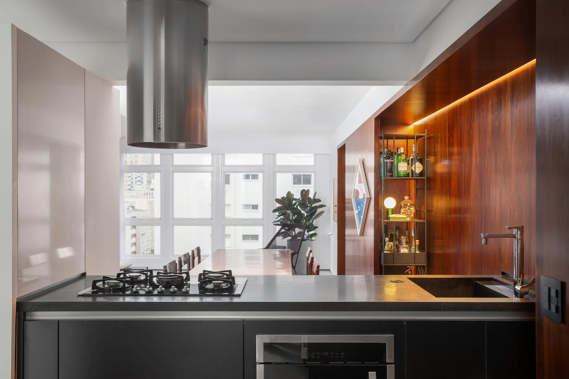 A modern kitchen island with a cooktop and sink stands in front of a warm wood-paneled wall with recessed bar shelving.