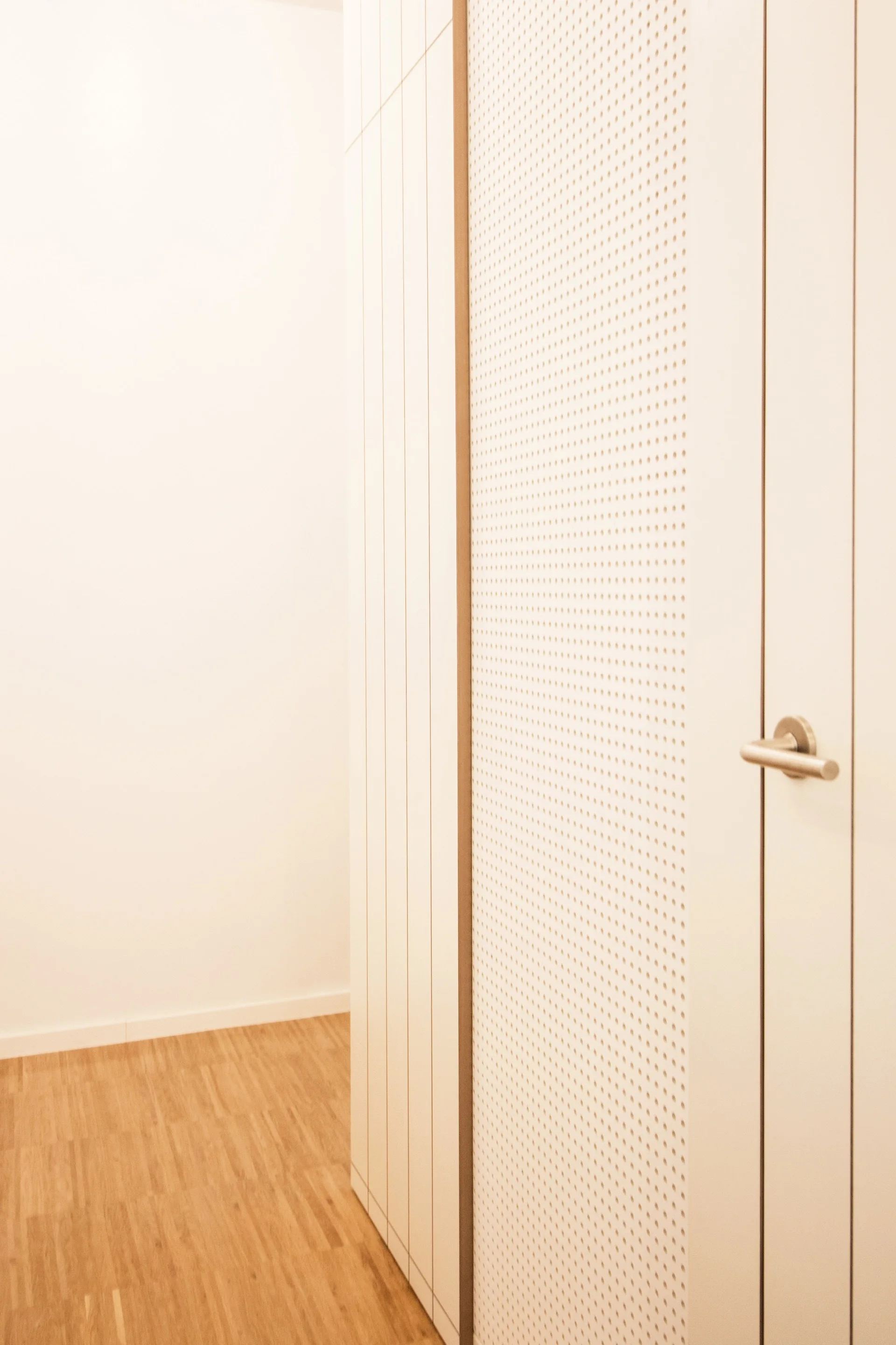 A close-up of an off-white perforated door with a silver lever handle next to vertical wall panels and a light wood floor.