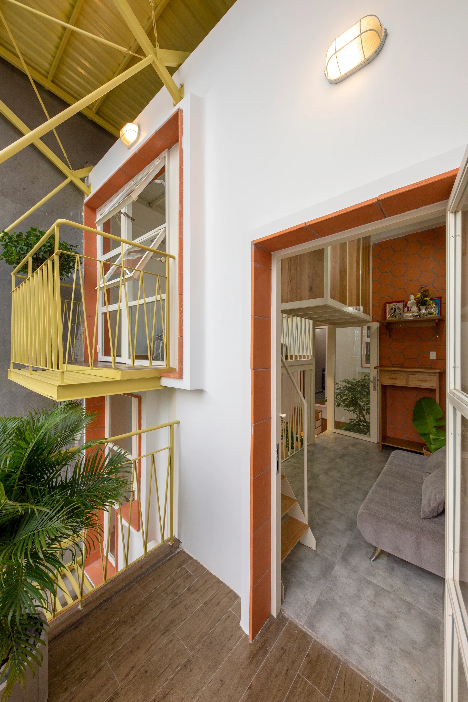 A split-level modern interior featuring a grey sofa, an indoor staircase, a bright yellow metal balcony railing, and orange tile door trim.
