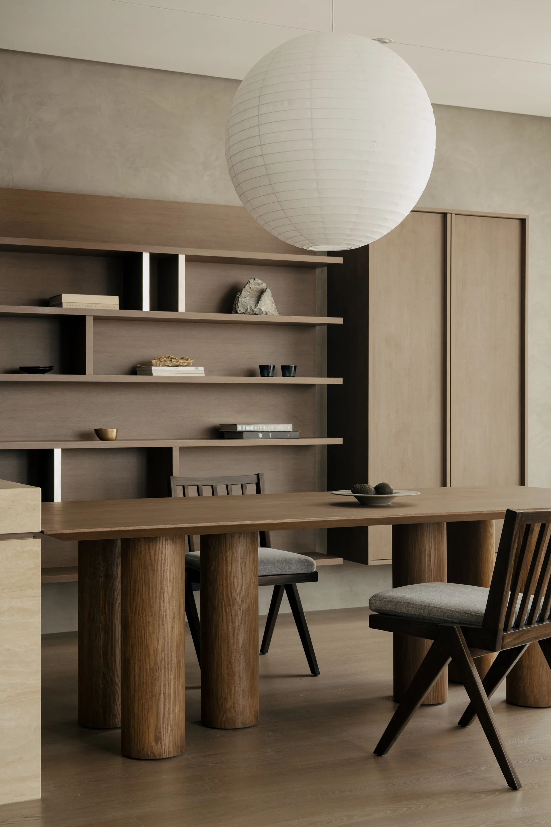 A minimalist dining room featuring a wooden table with thick cylindrical legs, slat-back chairs, built-in shelving, and a large white paper pendant light.