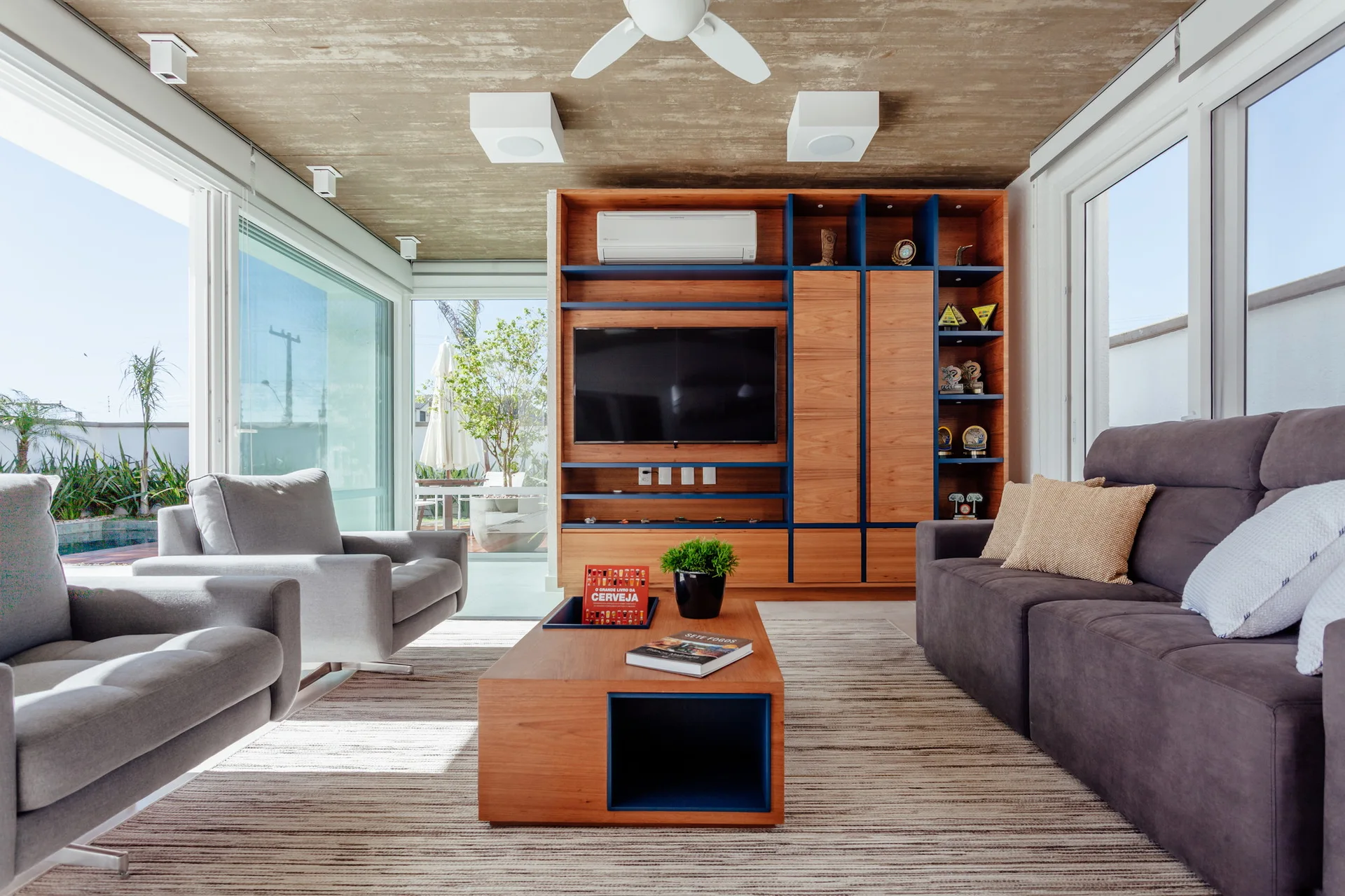 A modern living room features a grey sofa, two armchairs, a custom wood and blue media unit, and a textured concrete ceiling.