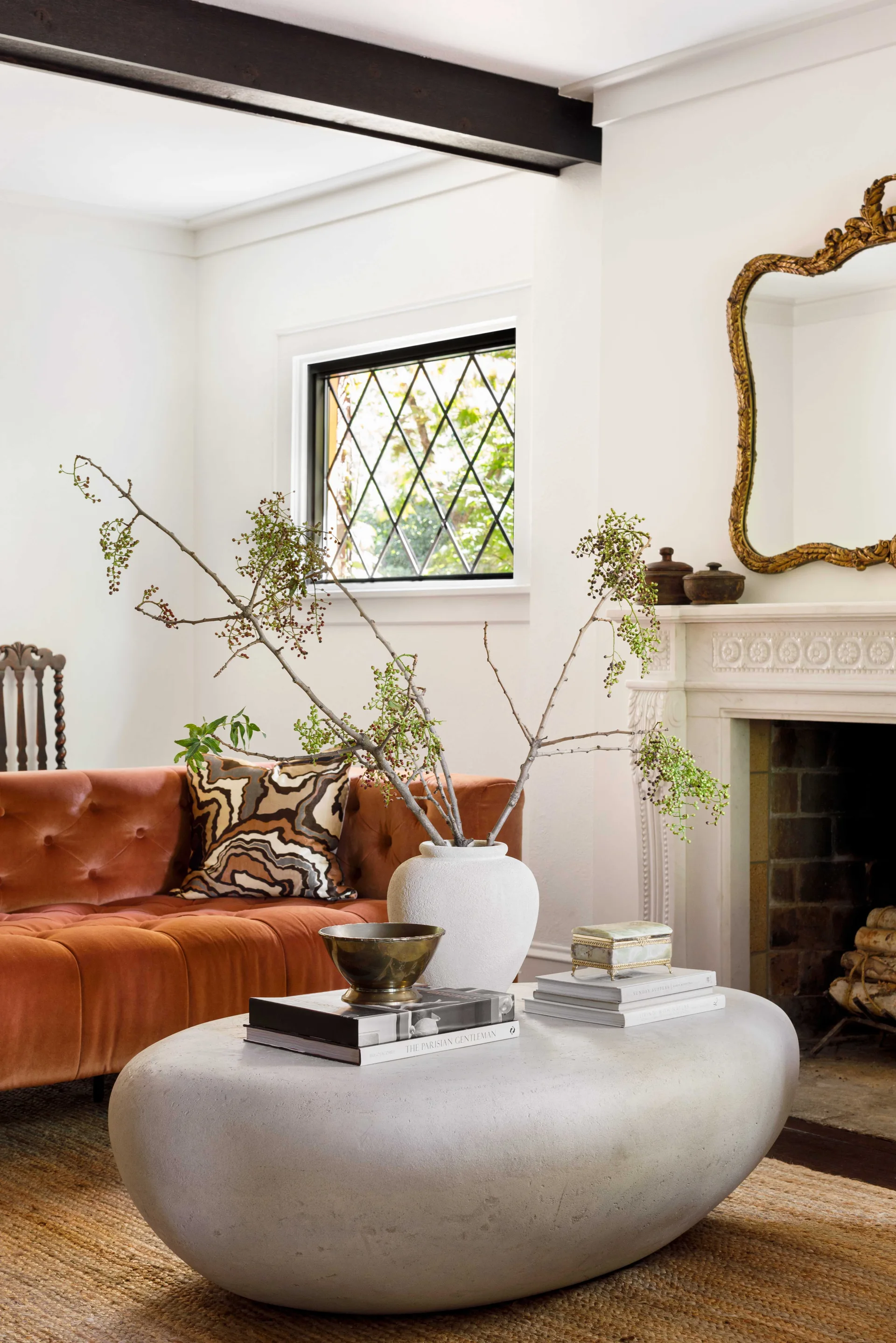 A living room features a tufted rust velvet sofa, a low pebble-shaped stone coffee table with branches in a white vase, and an ornate fireplace.