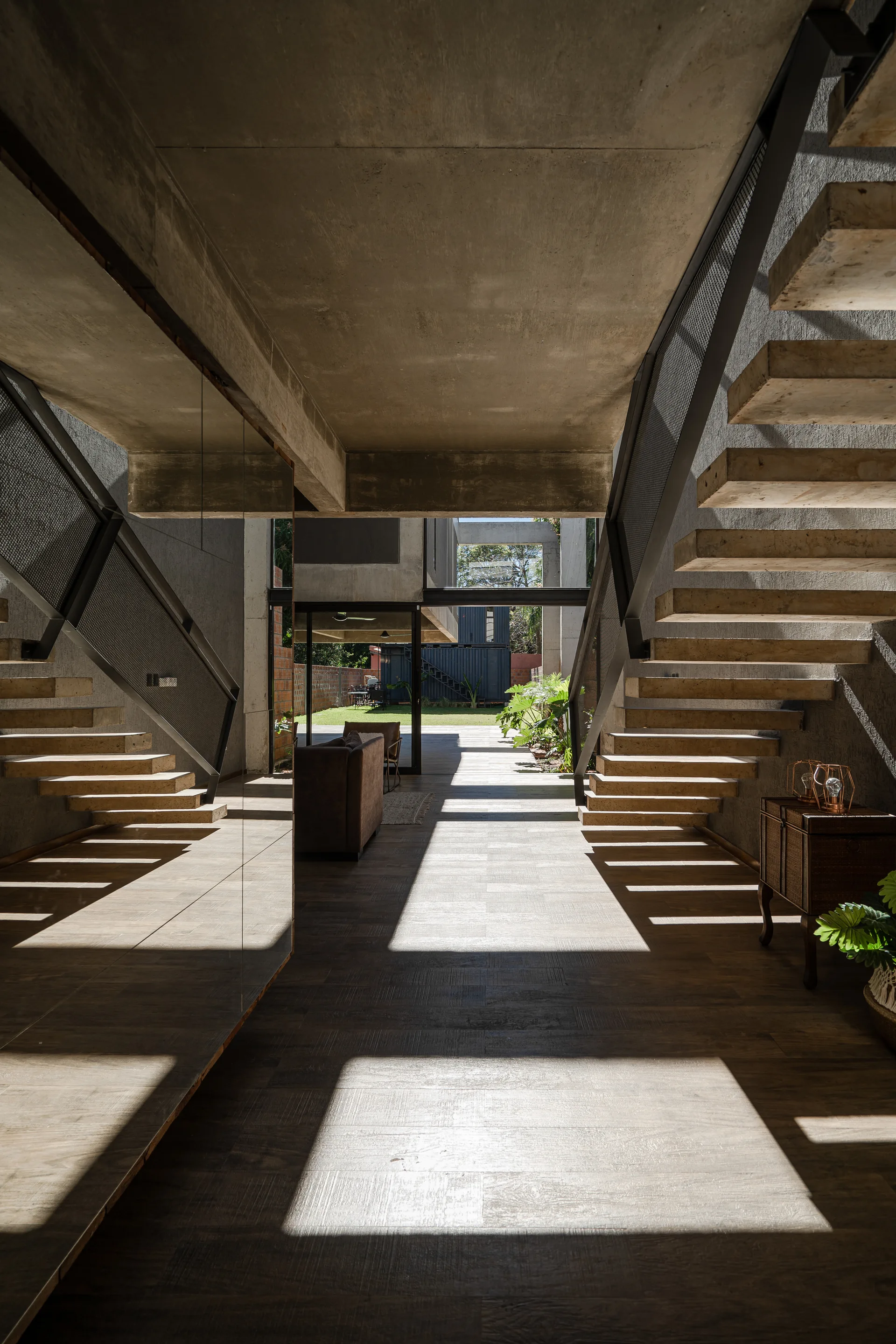 An industrial hallway features raw concrete walls, a floating cantilevered staircase, and a large mirror reflecting the stairs under dramatic natural light.