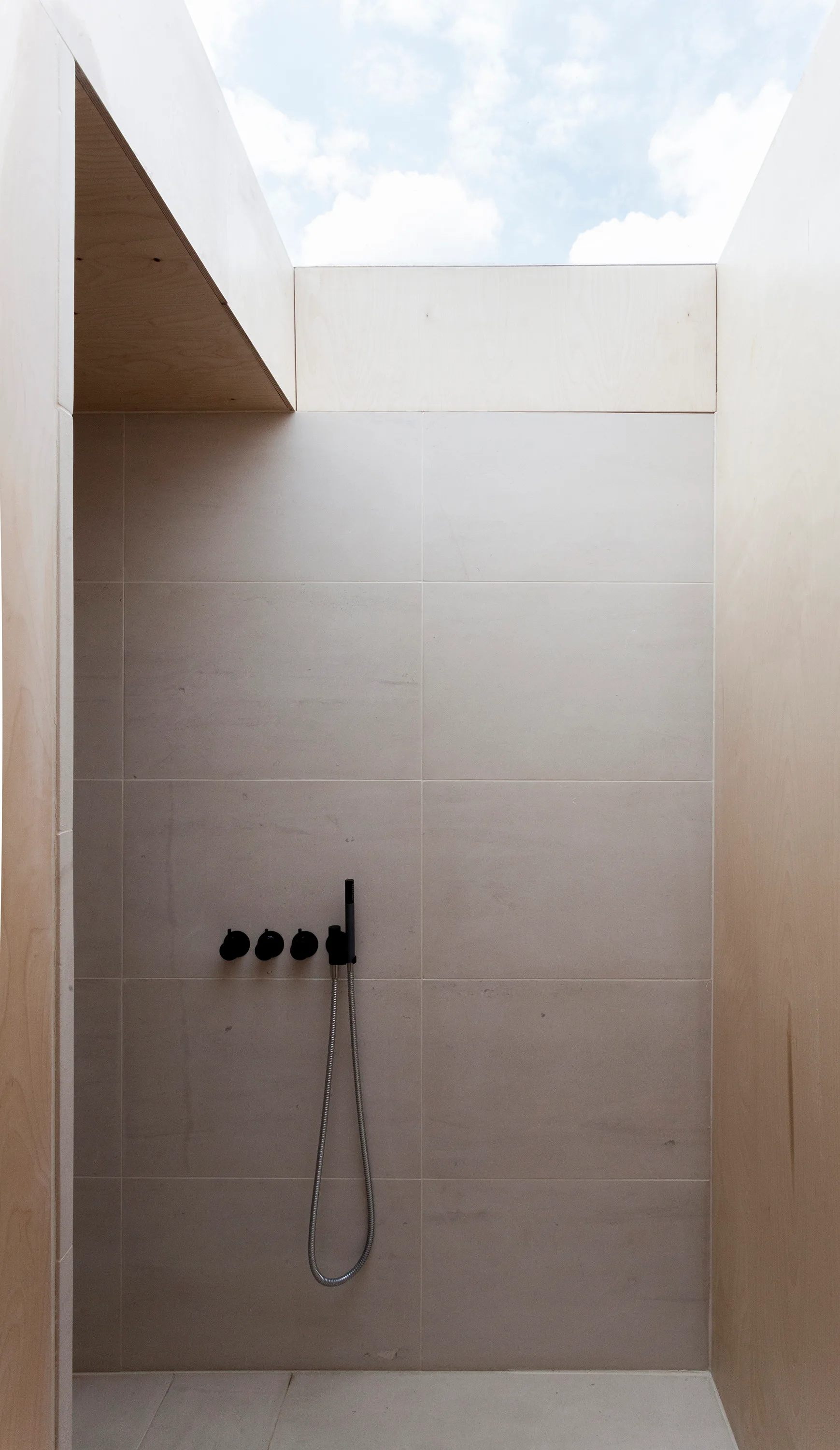 A minimalist shower featuring large beige tiles, black matte fixtures, light wood side panels, and a large open skylight showing the sky.