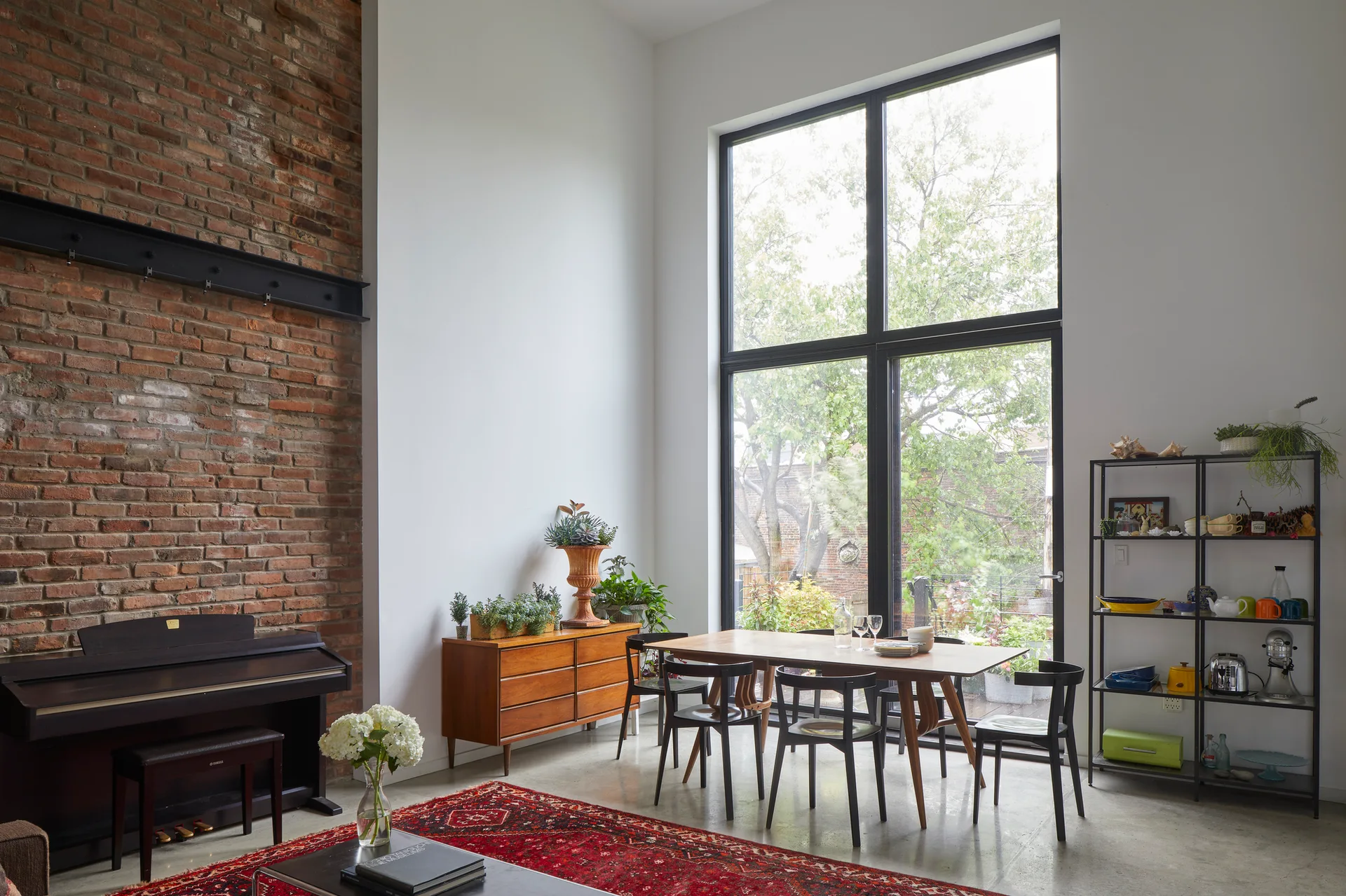 A dining room features a wooden table with black chairs, a large garden-facing window, an exposed brick wall, and a red patterned rug.