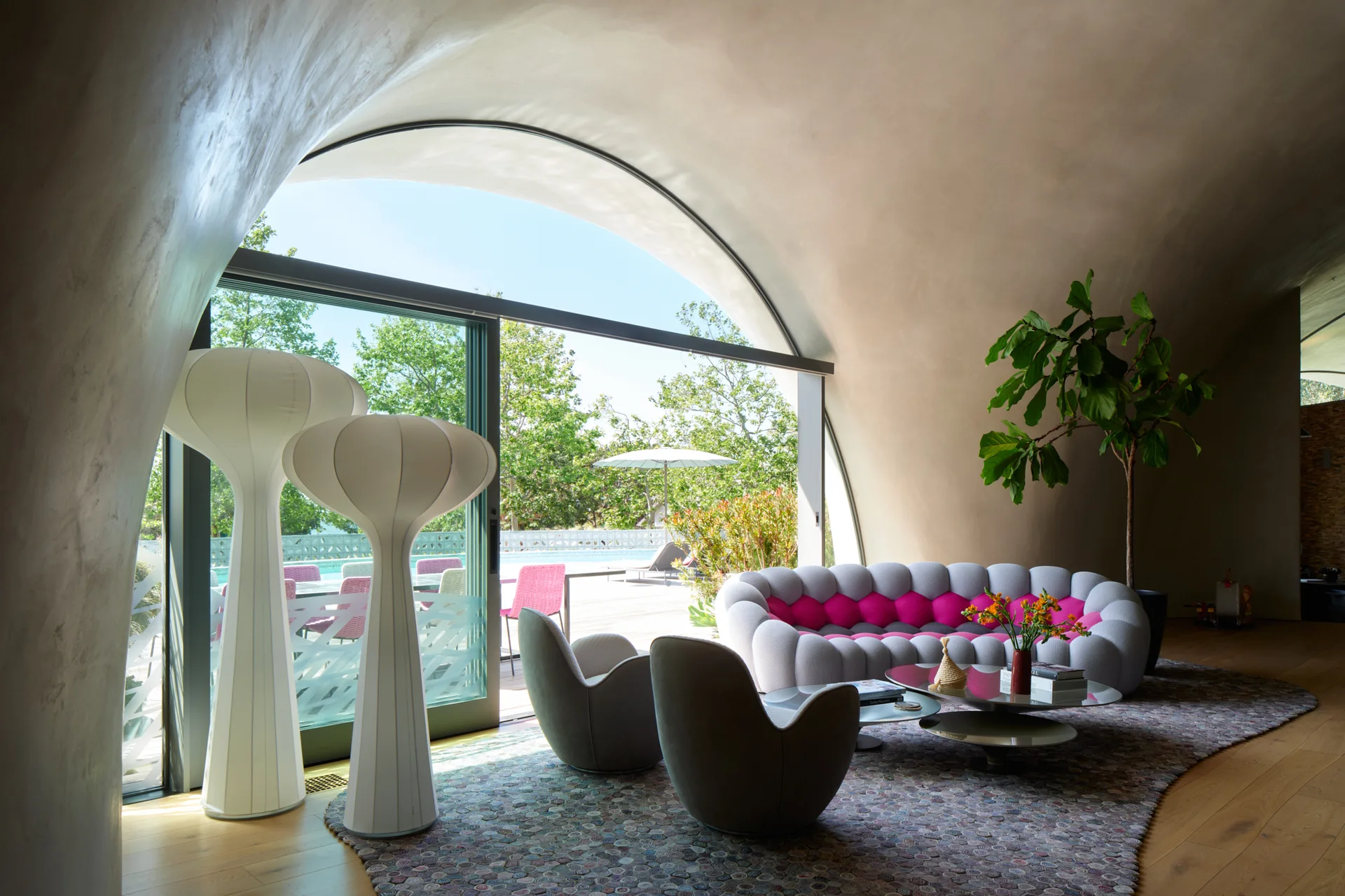A modern living room under a curved vaulted ceiling features a grey and magenta tufted sofa, two white floor lamps, and a large sliding glass door.