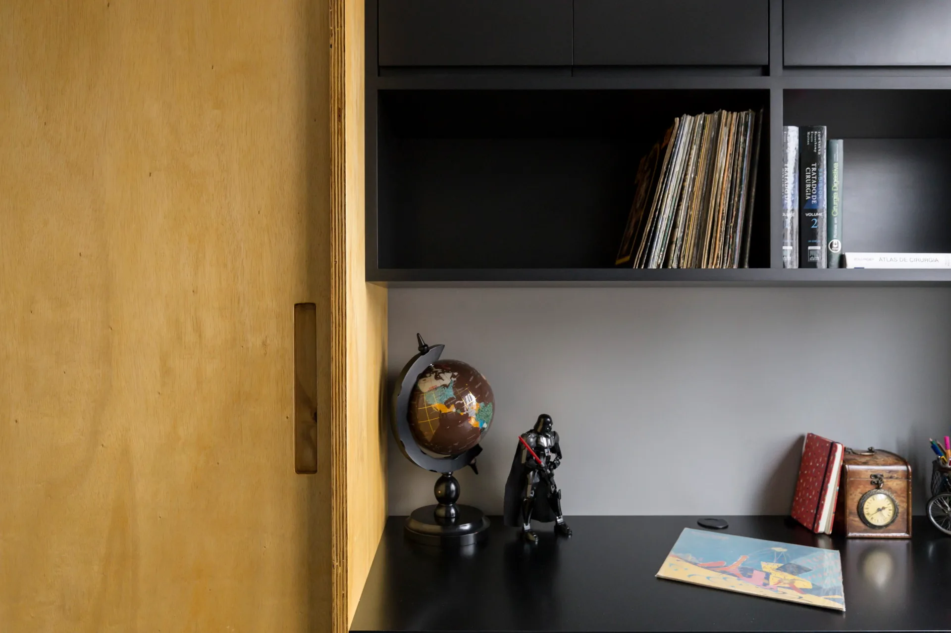 A built-in home office workspace features a matte black desk and matching overhead shelving next to a light plywood cabinet.