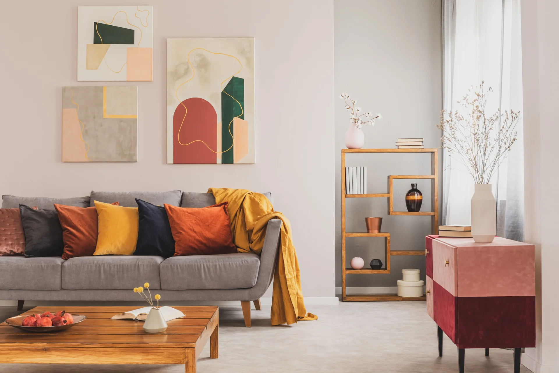 A grey fabric sofa features colorful velvet throw pillows next to an asymmetrical wooden bookcase and a pink and burgundy cabinet.