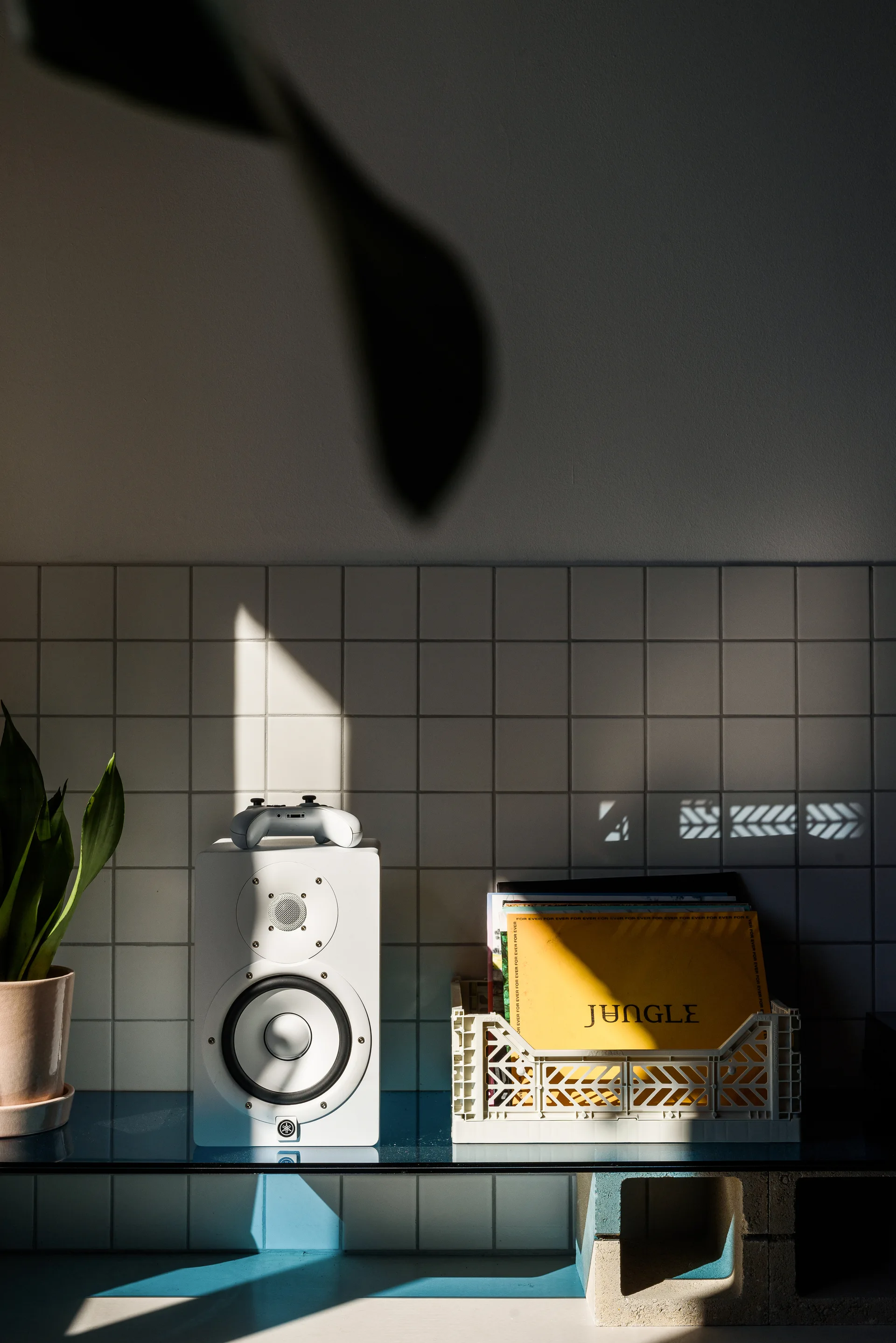 A white studio speaker, a plastic crate of vinyl records, and a potted plant sit on a glass shelf supported by cinder blocks against a square-tiled wall.