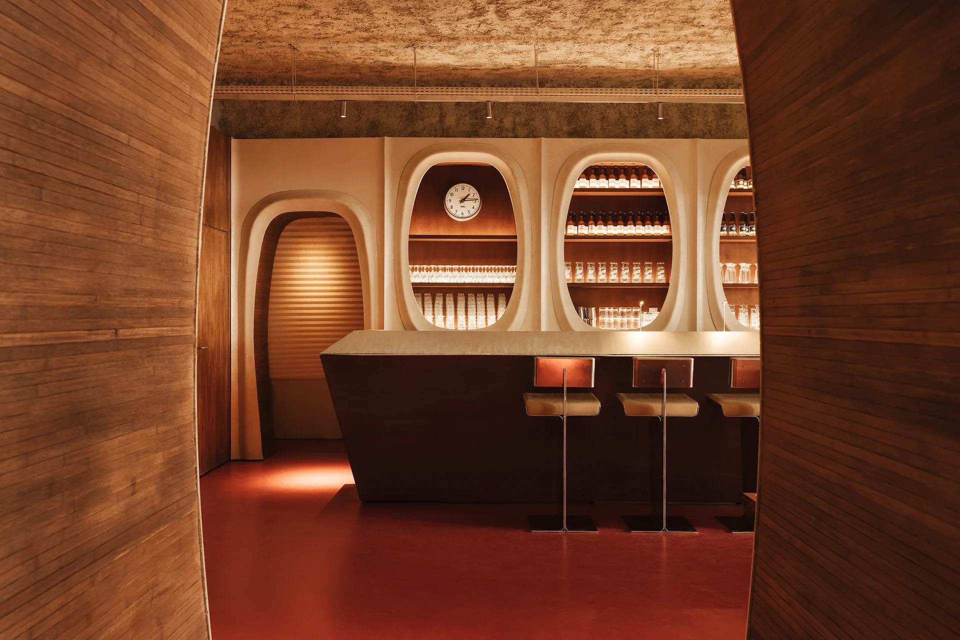 A retro-style bar interior featuring wood-slatted walls, three metal-framed stools, a dark counter, and backlit capsule-shaped shelving niches.