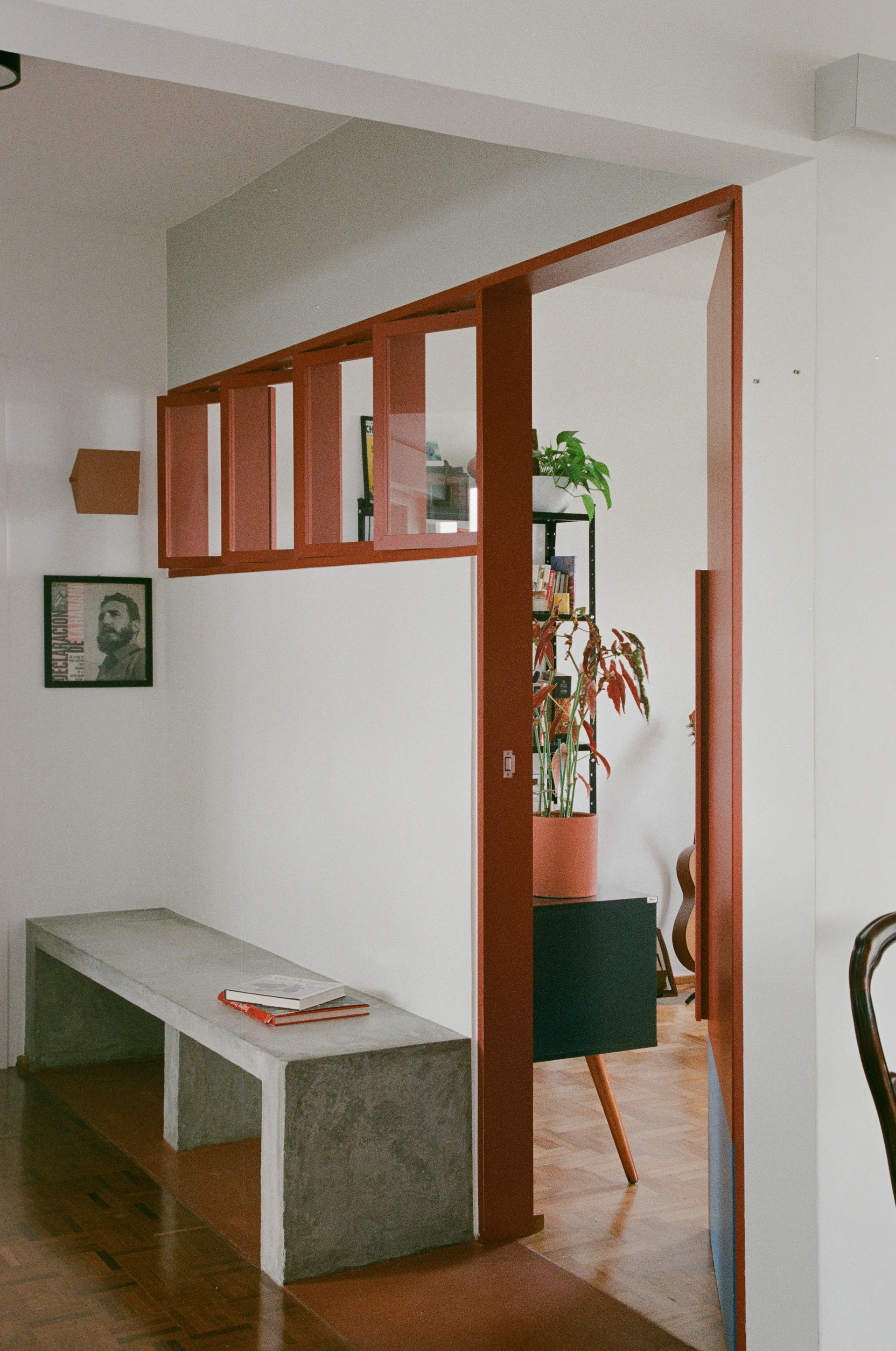 A minimalist entryway featuring a gray concrete bench against a white wall and a terracotta-painted door frame with a glass transom window.