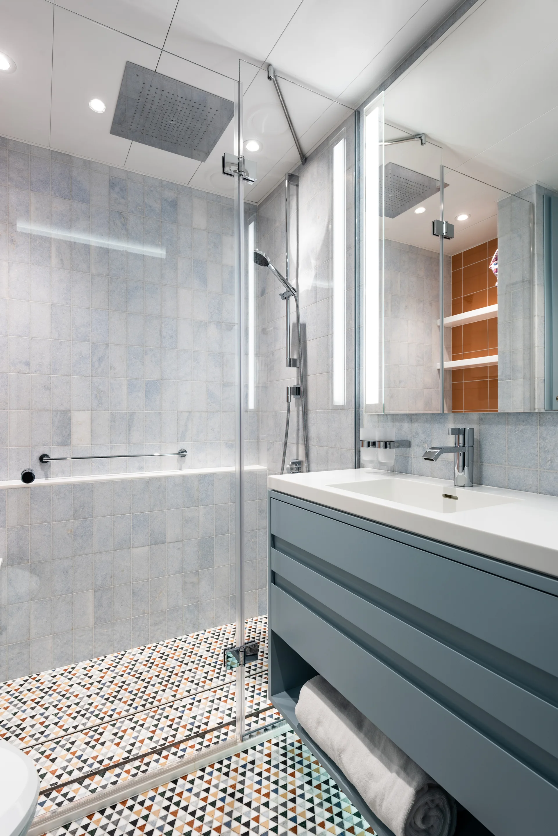 A modern bathroom featuring a blue floating vanity, light blue wall tiles, and a colorful geometric triangular tile floor.