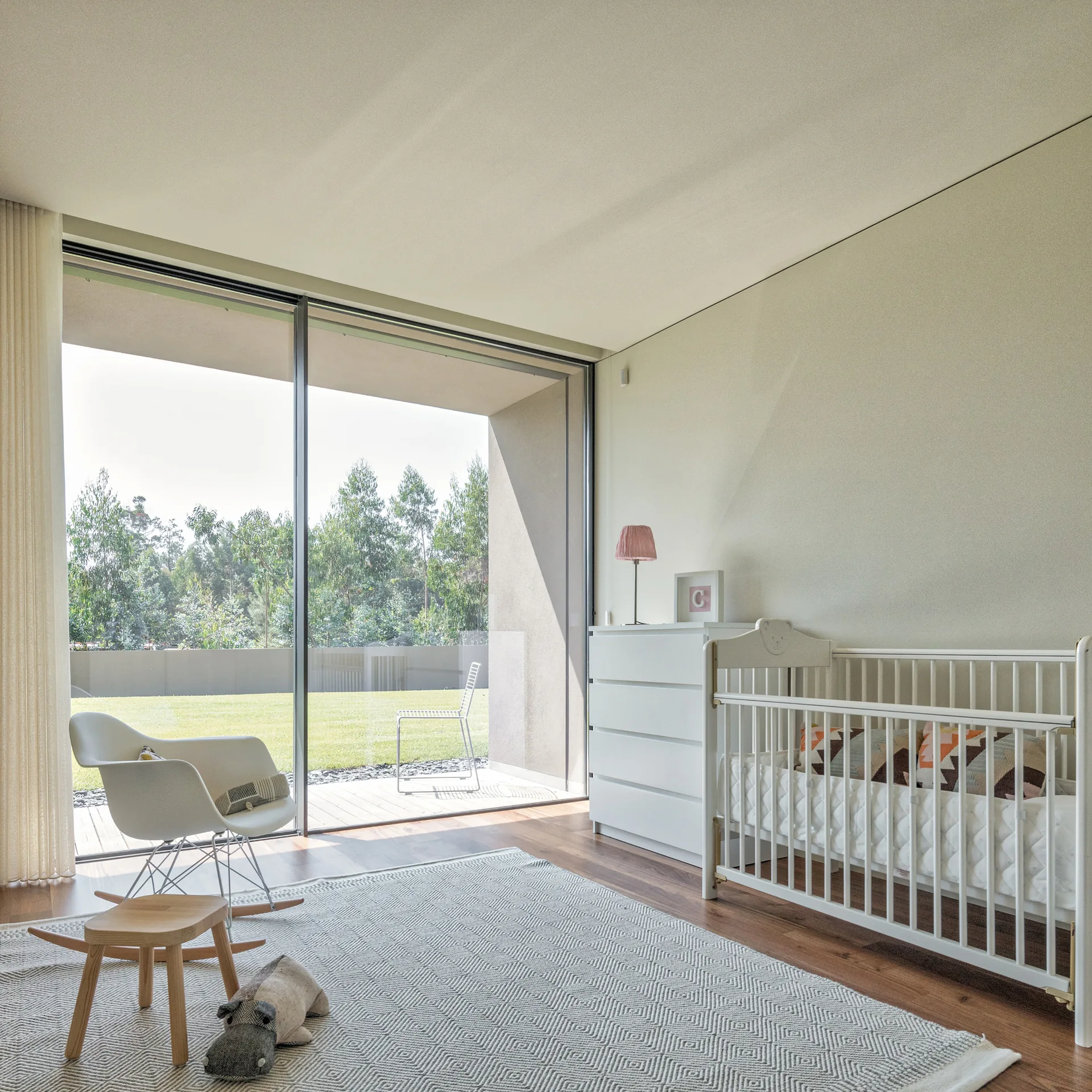 A white baby crib and mid-century modern rocking chair sit on a geometric rug in a bright nursery with large glass doors opening to a grassy lawn.