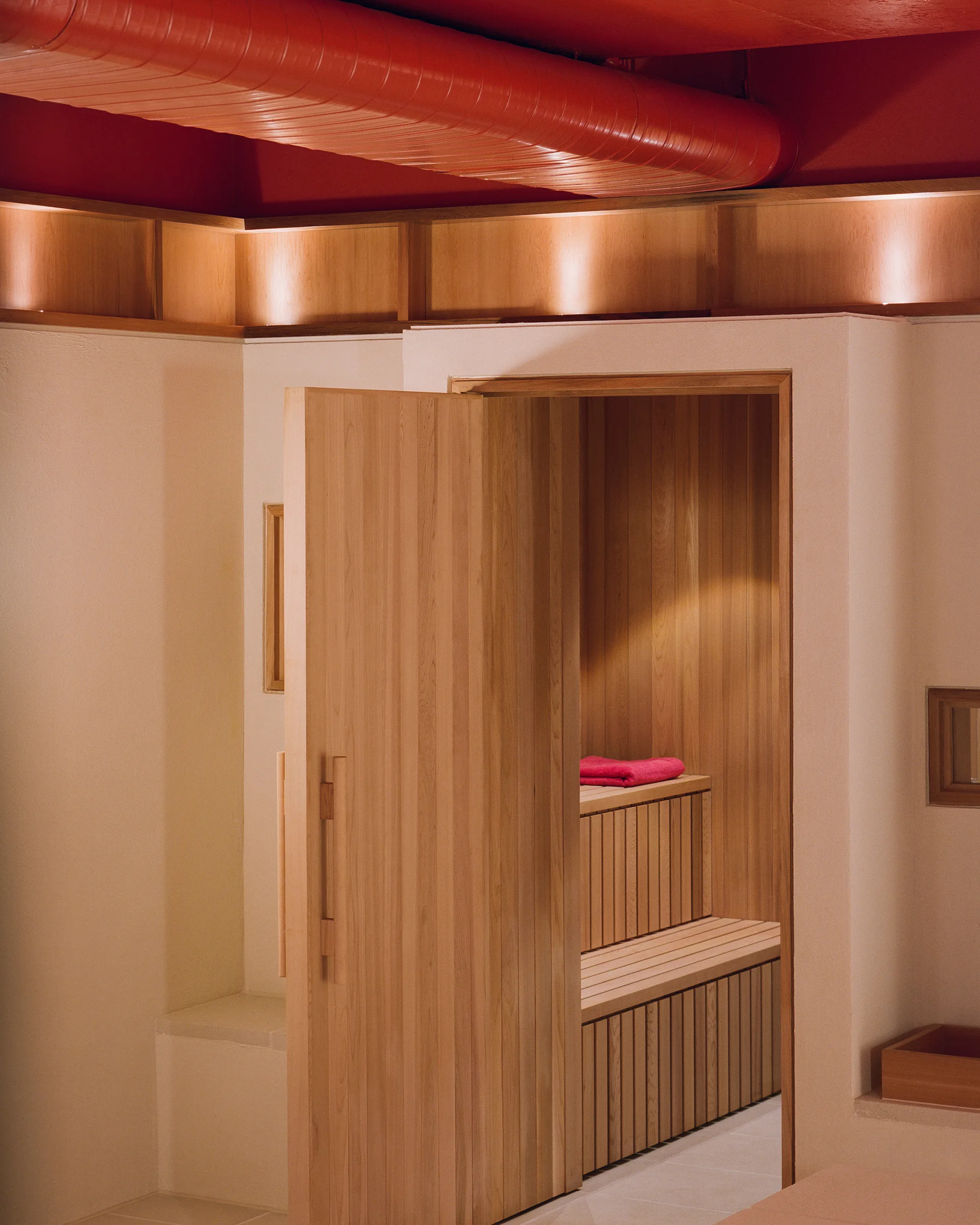 An interior view of a wooden sauna featuring tiered benches and vertical paneling under a red painted ceiling with an exposed matching duct.
