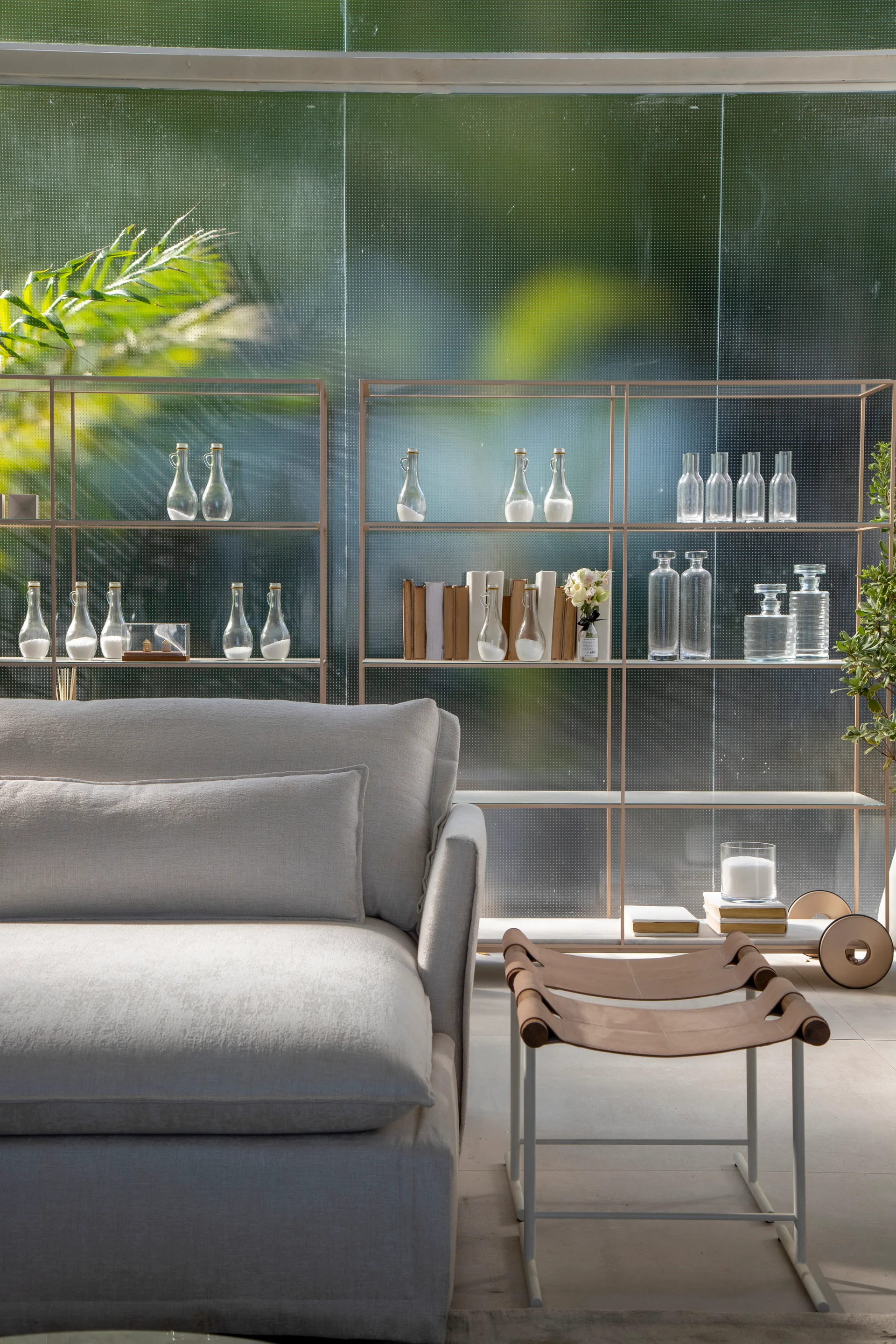A light grey upholstered sofa and a tan leather sling stool sit before a metal display shelf styled with bottles against a dotted glass wall.