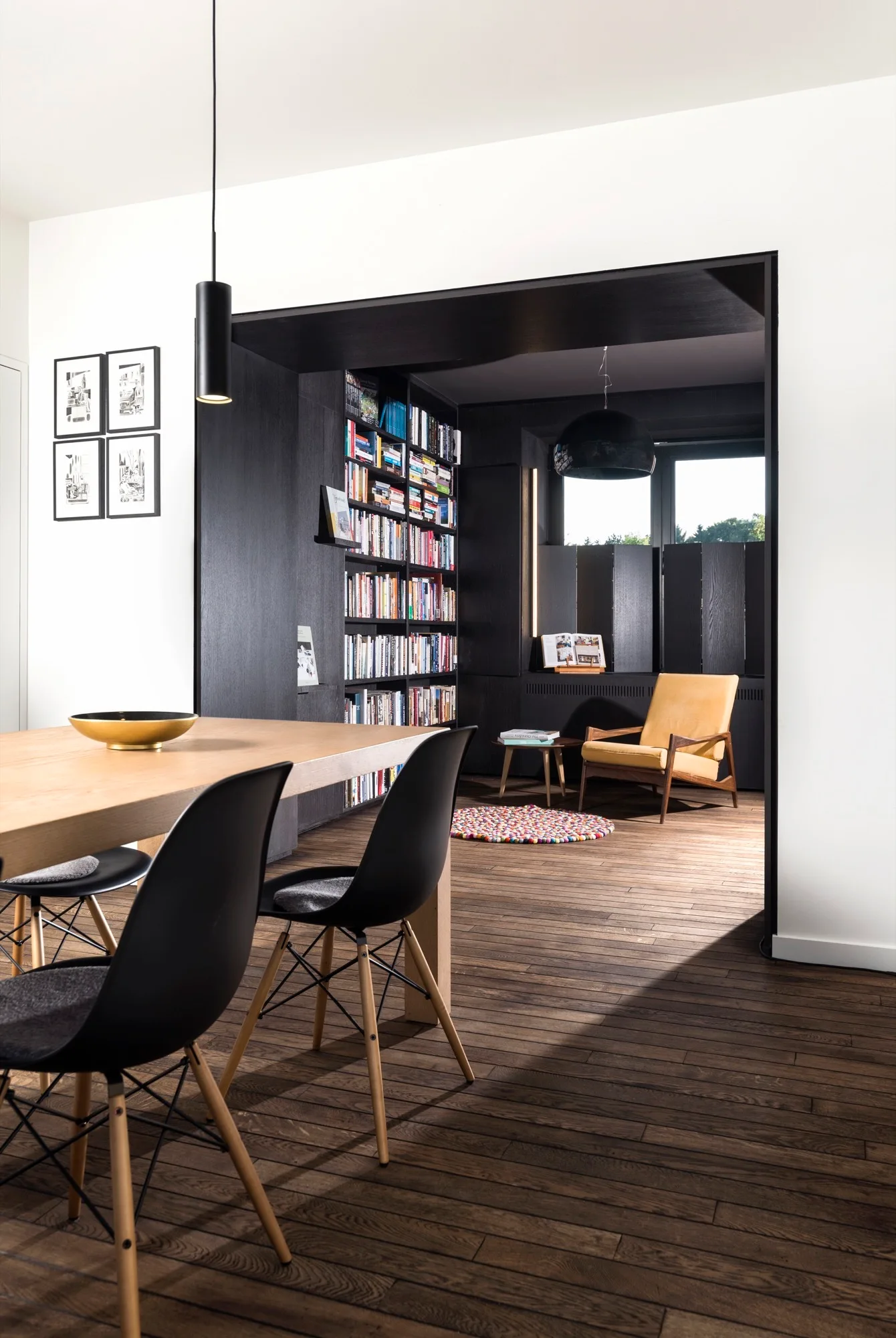A wood dining table with black chairs sits next to a dark wood archway opening into a reading room with a tall built-in bookcase and a yellow armchair.