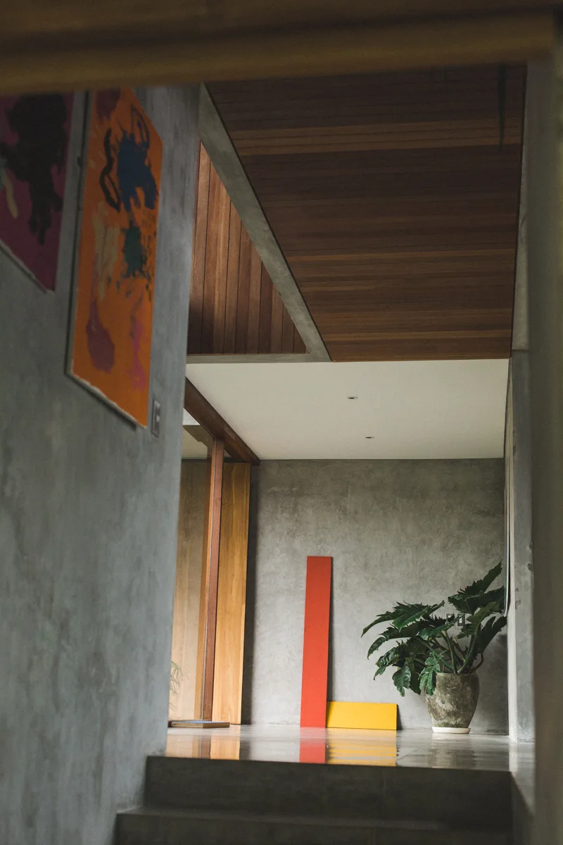 A minimalist gray concrete hallway features a wood slatted ceiling, polished floors, and vibrant leaning art panels near a potted plant.