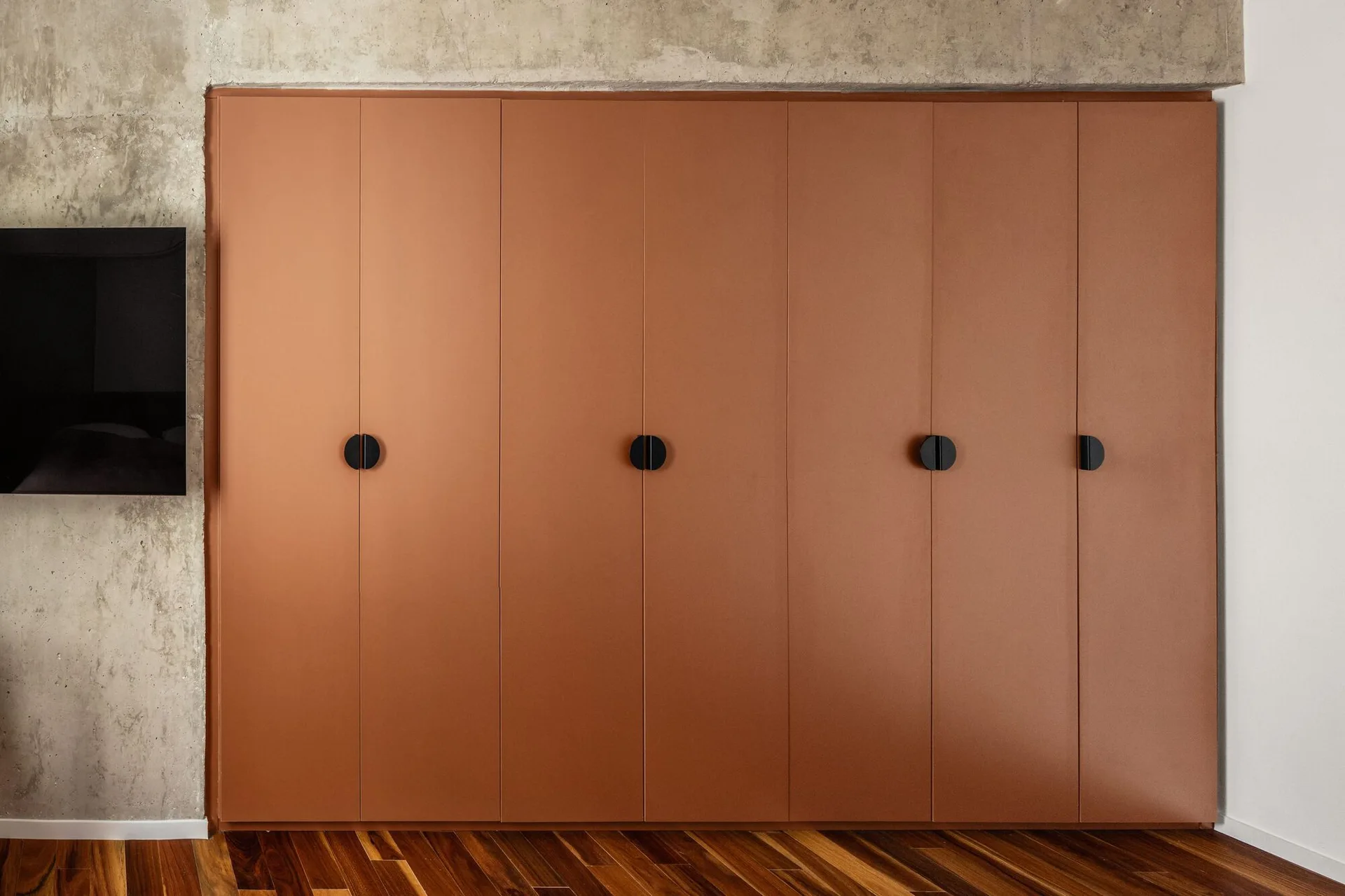A modern terracotta closet with round black handles is built against a raw concrete wall above a dark hardwood floor.