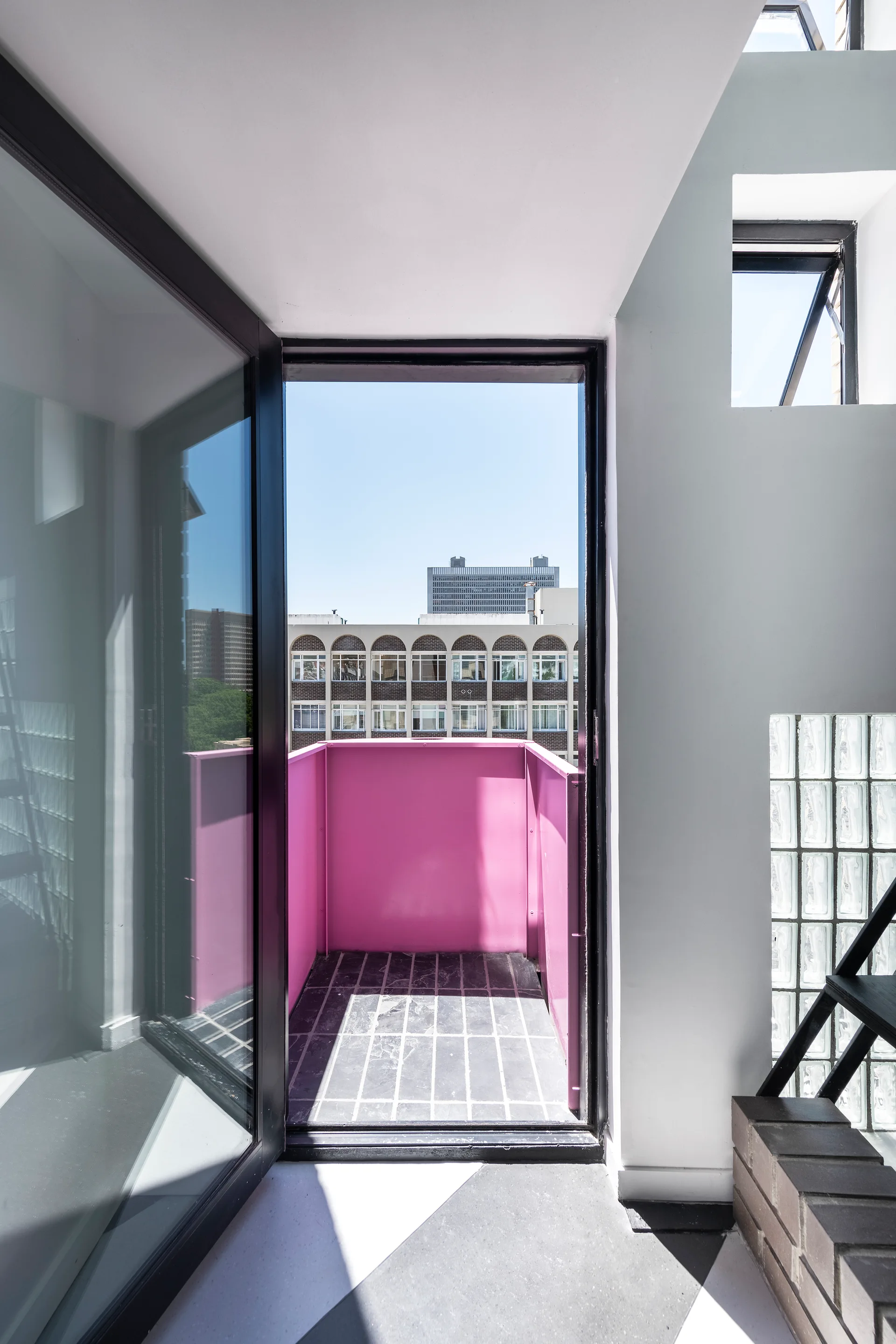 A view from a white minimalist interior through an open doorway onto a small balcony with a bright pink metal railing and grey paved floor.