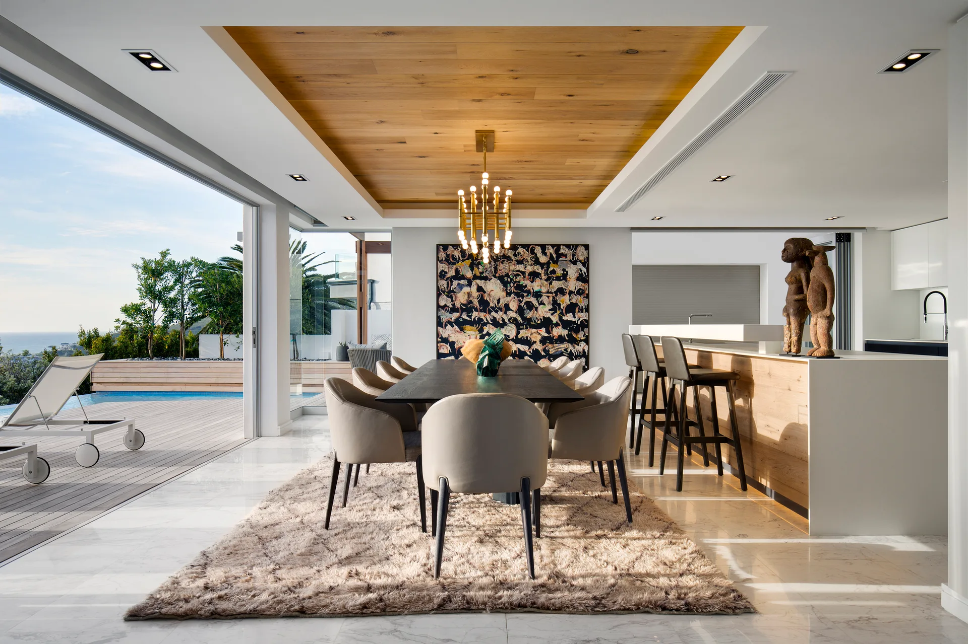 A contemporary dining room with a dark wood table under a recessed wood ceiling panel, opening to an outdoor pool deck through sliding glass doors.