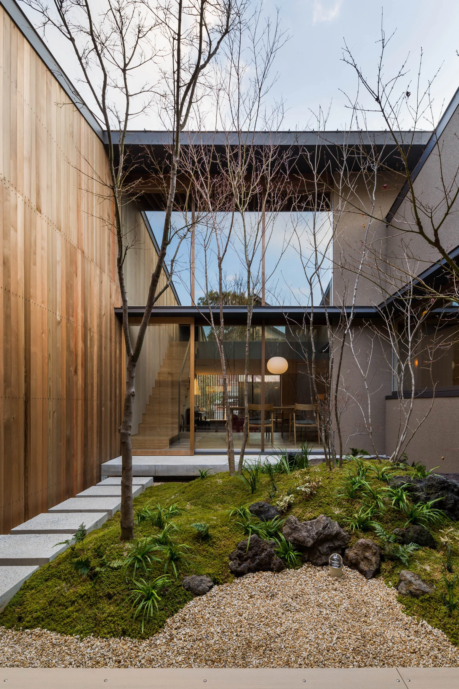 A modern residential courtyard features a landscaped moss garden with young trees, viewed through large glass walls showing a wooden staircase inside.