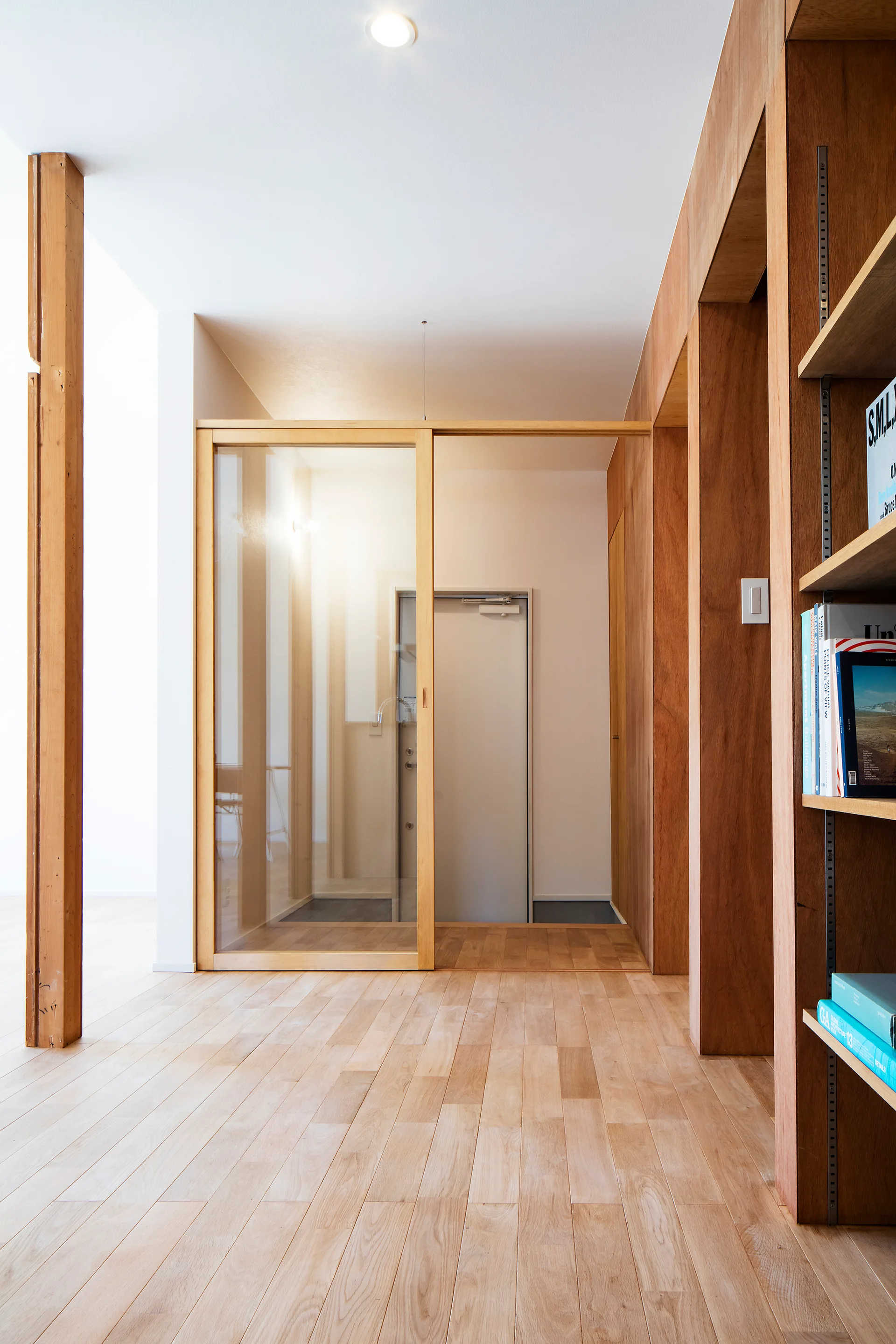 A minimalist entryway featuring light wood flooring, warm wood paneled walls, a sliding glass partition door, and integrated wooden shelving.