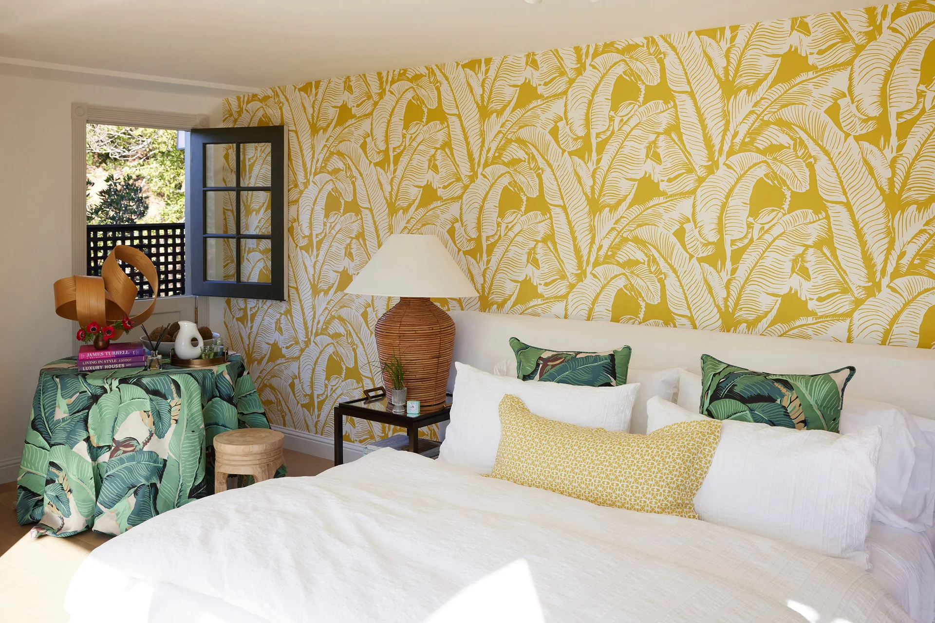 A bright bedroom featuring bold yellow leaf wallpaper, a white bed with patterned throw pillows, and a large woven table lamp.