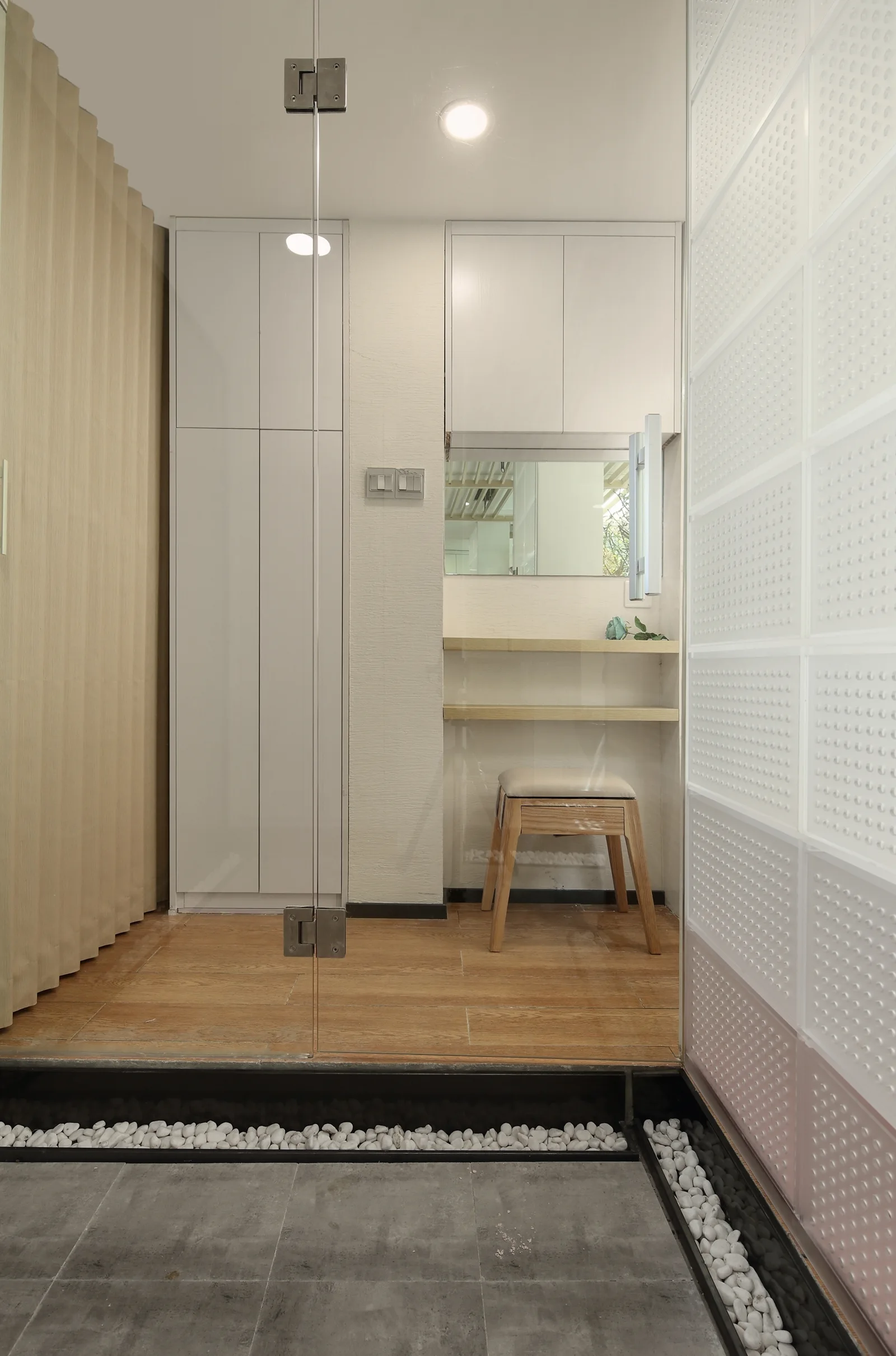 A minimalist dressing area features built-in white cabinets, a floating wood vanity shelf with a stool, and a textured glass block partition wall.