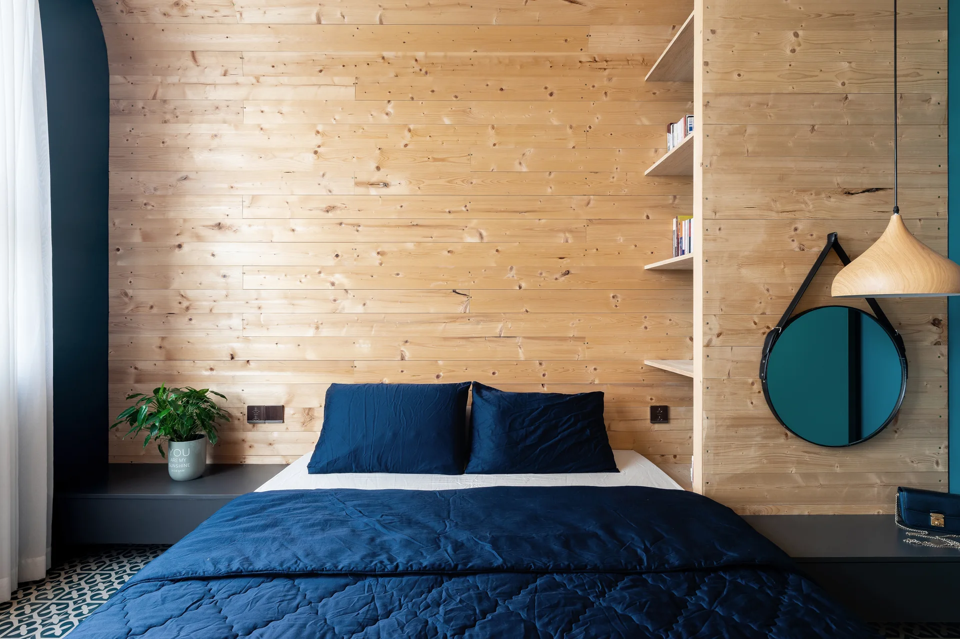 A bed with navy blue bedding sits against a horizontal light wood plank wall featuring integrated shelves and dark floating bedside ledges.