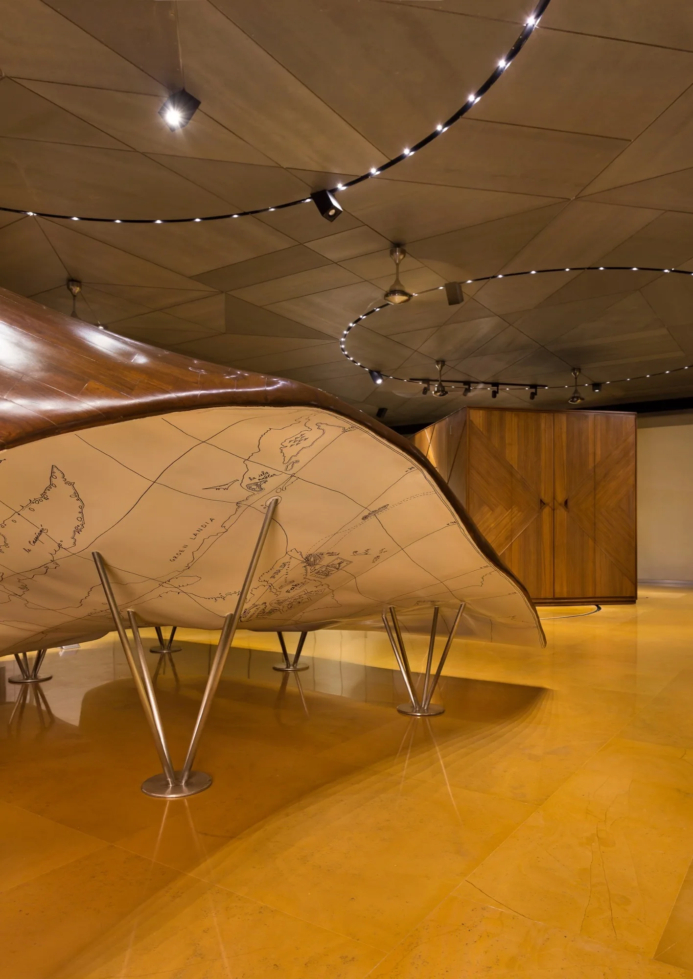 A sculptural wood table featuring an illustrated map on its underside stands on a glossy yellow floor beside a geometric-patterned cabinet.