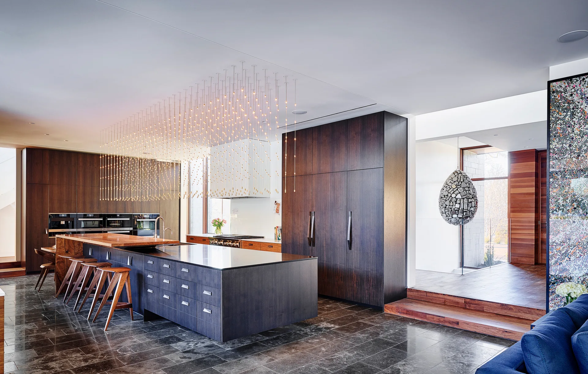 A modern kitchen with dark wood cabinetry, a large central island with stools, and an extensive cloud-like pendant light installation.