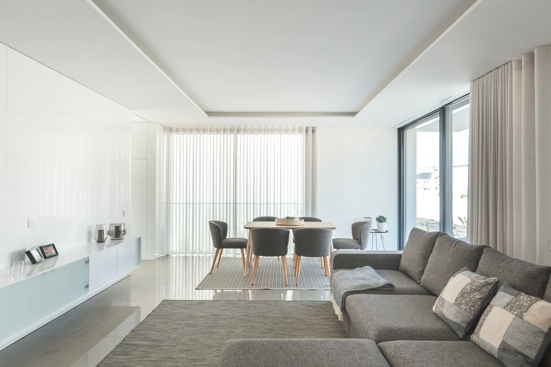 A bright open-concept living and dining room with a grey sectional sofa, two area rugs, a wooden dining table with chairs, and glossy white built-in cabinets.