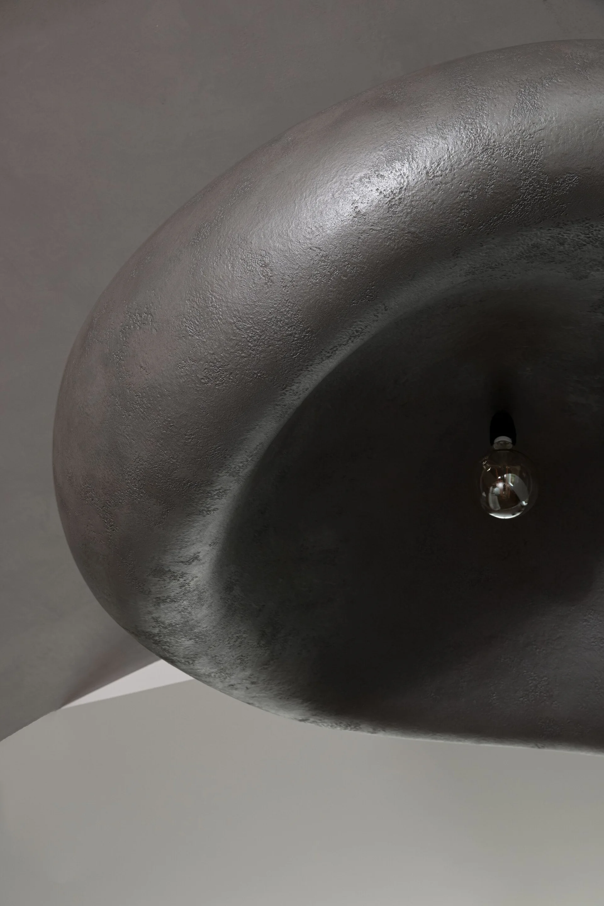 A close-up view of a large sculptural pendant light with a textured dark gray plaster finish and a small exposed light bulb hanging beneath it.