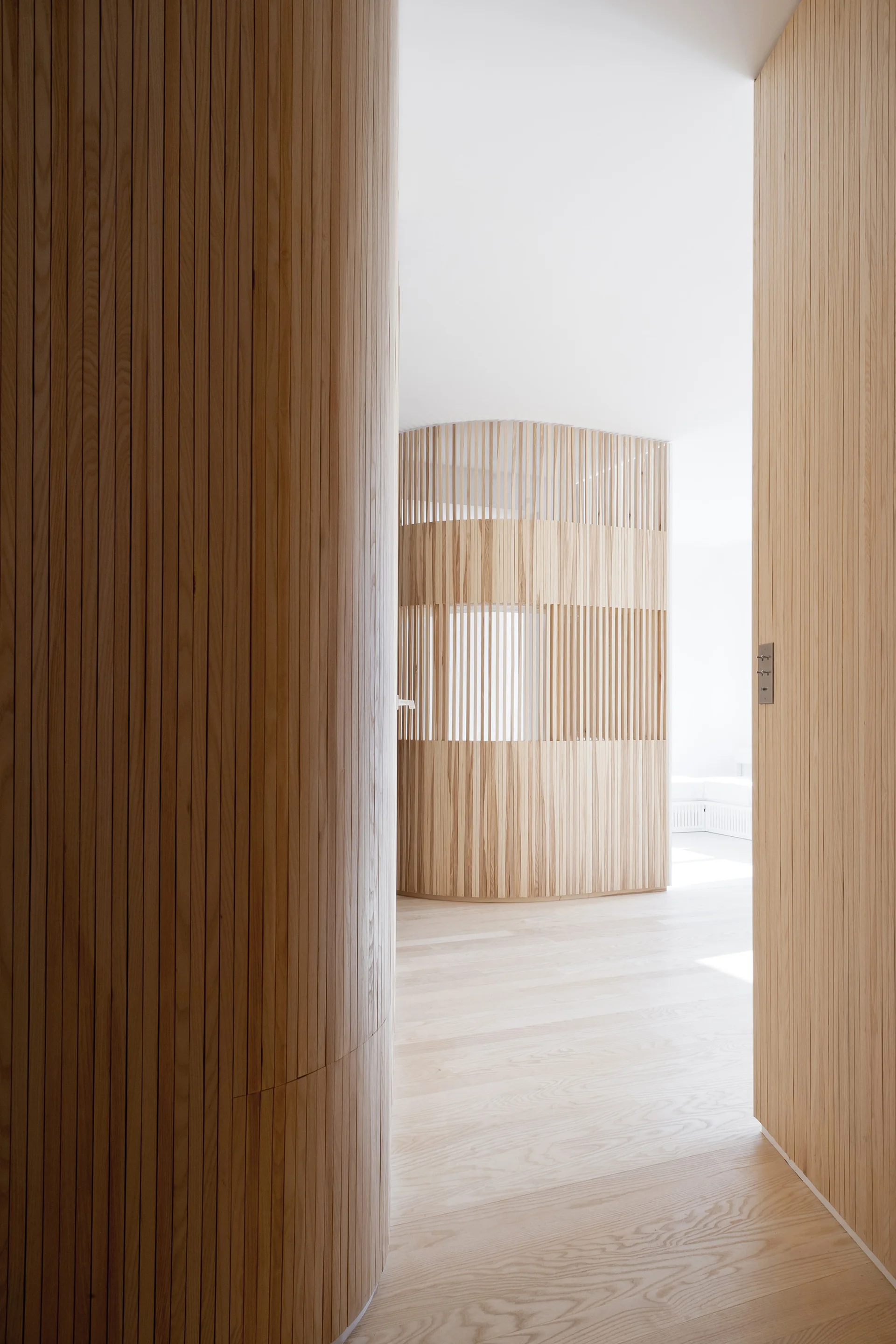A minimalist hallway featuring light wood flooring and curved walls clad in vertical wooden slats that create a semi-private partition.