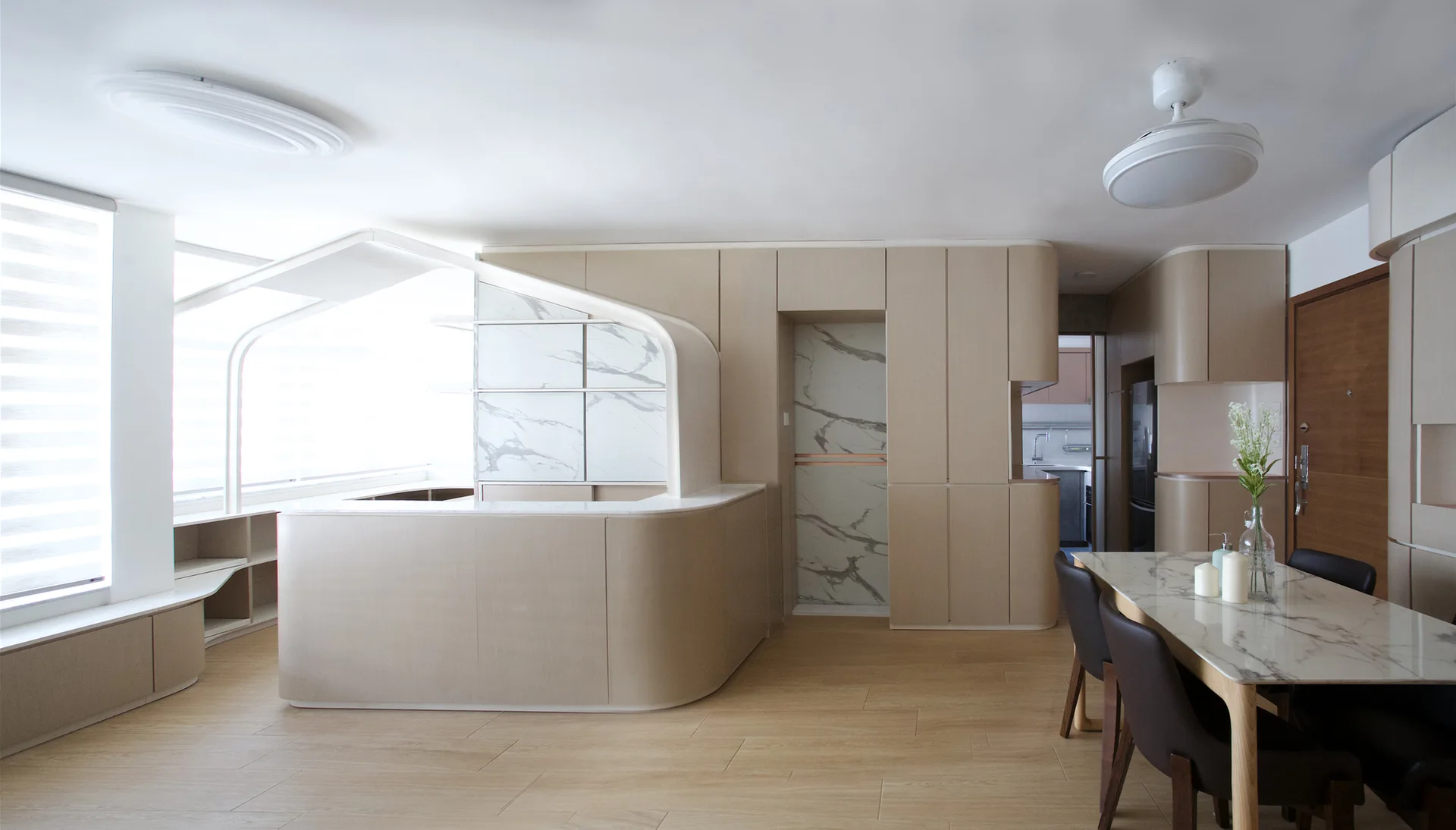 A modern open-plan dining area featuring a marble-topped table, curved beige cabinetry, and light wood flooring under a white ceiling.