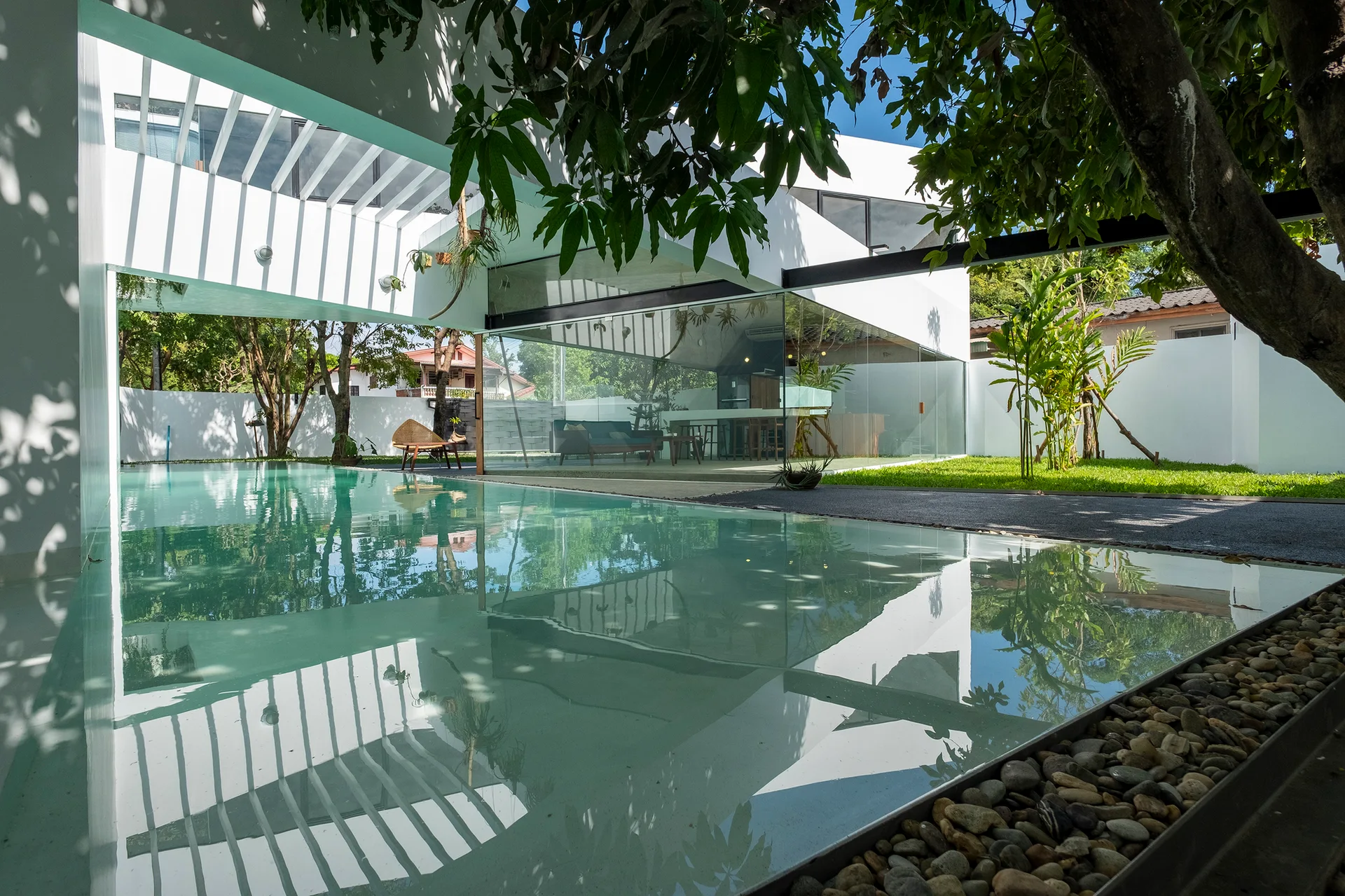 A modern white house features a large swimming pool, slatted pergola overhead, and expansive glass walls showing the interior living area.