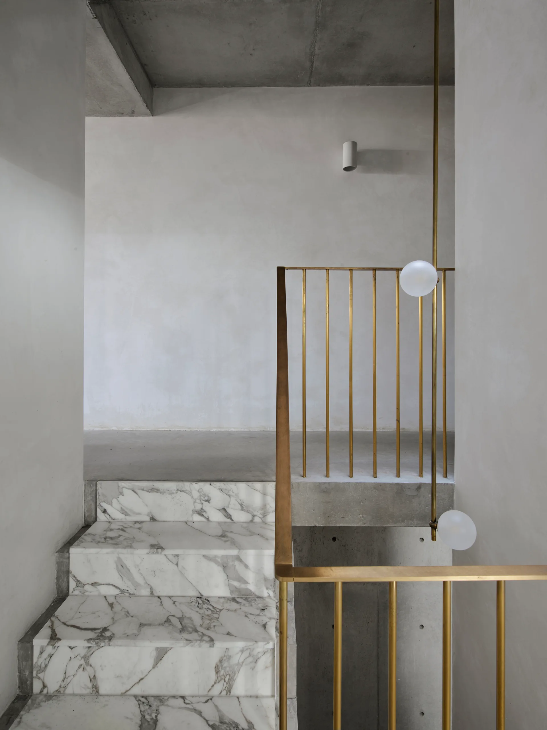 A modern staircase features veined white marble steps, vertical brass balusters, concrete walls, and minimalist globe pendant lights.