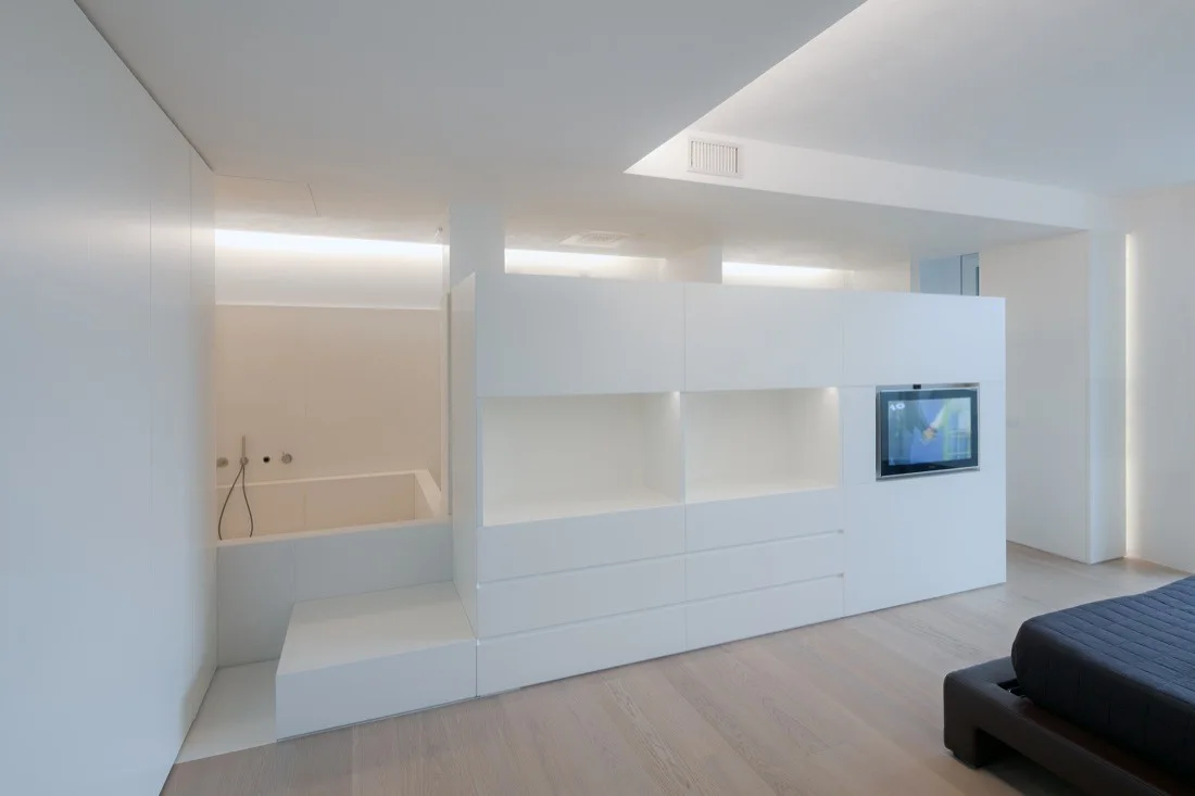 A minimalist bedroom features a large white partition wall with built-in storage and a TV, separating the bed from a recessed white bathtub.
